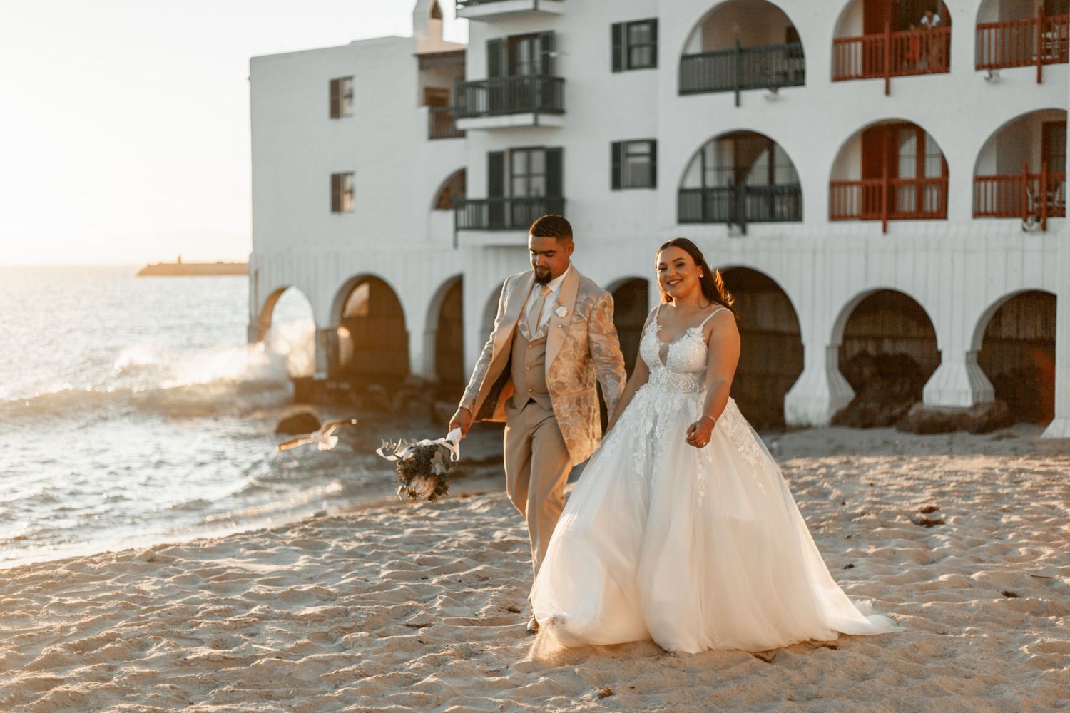 A couple walks along a beach at sunset in front of a Mediterranean-style building with arched windows and white walls.