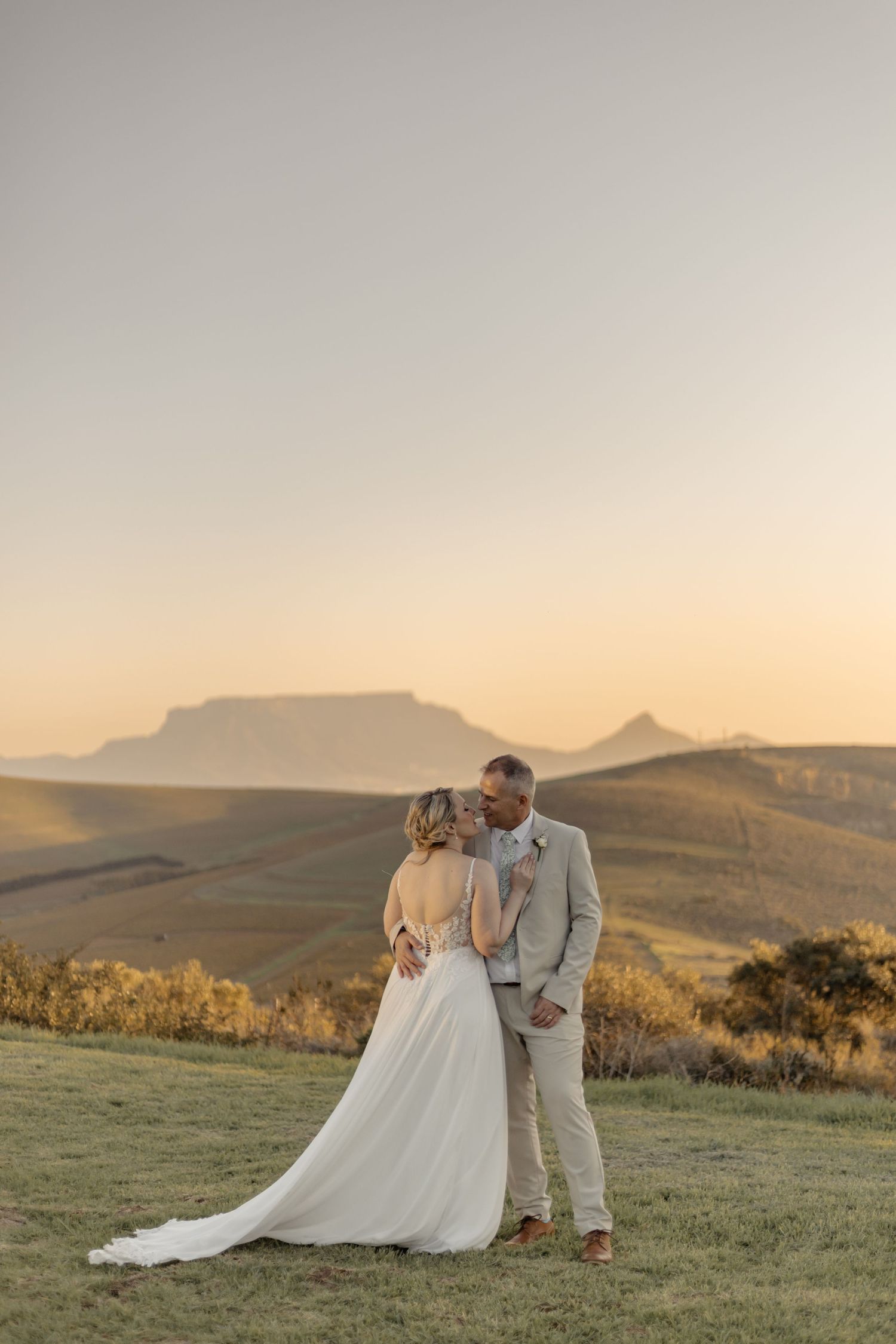 A couple in wedding attire embraces on a hillside at sunset with mountain silhouettes in the background.