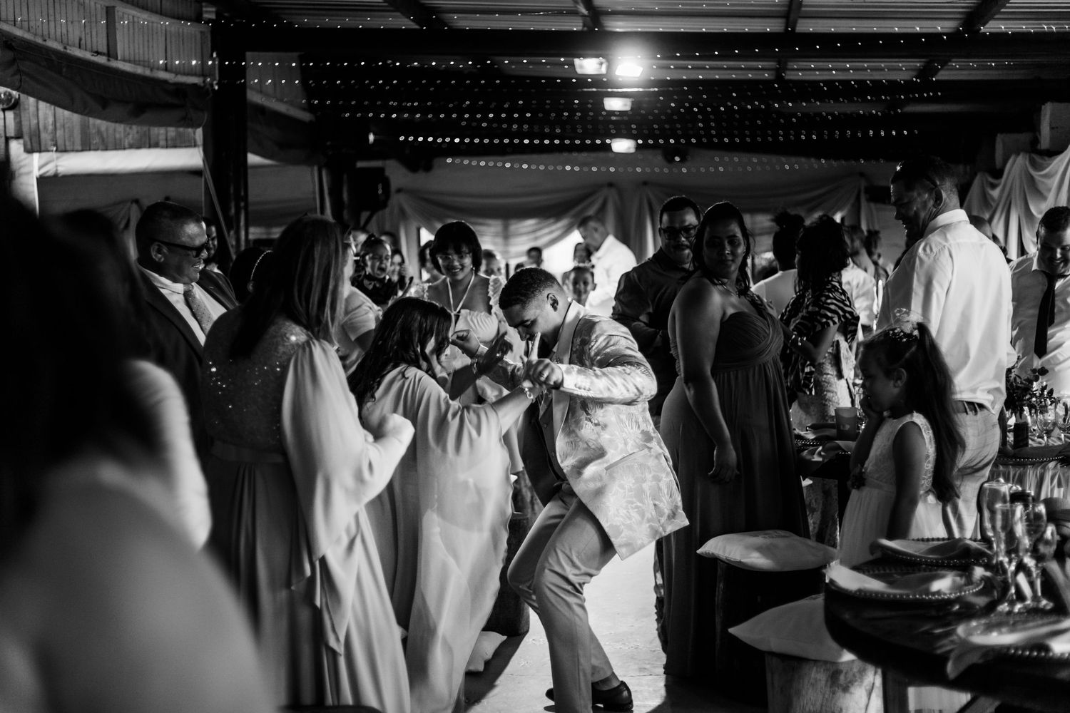 Black and white photo of people dancing and celebrating at a festive indoor party with hanging lights.