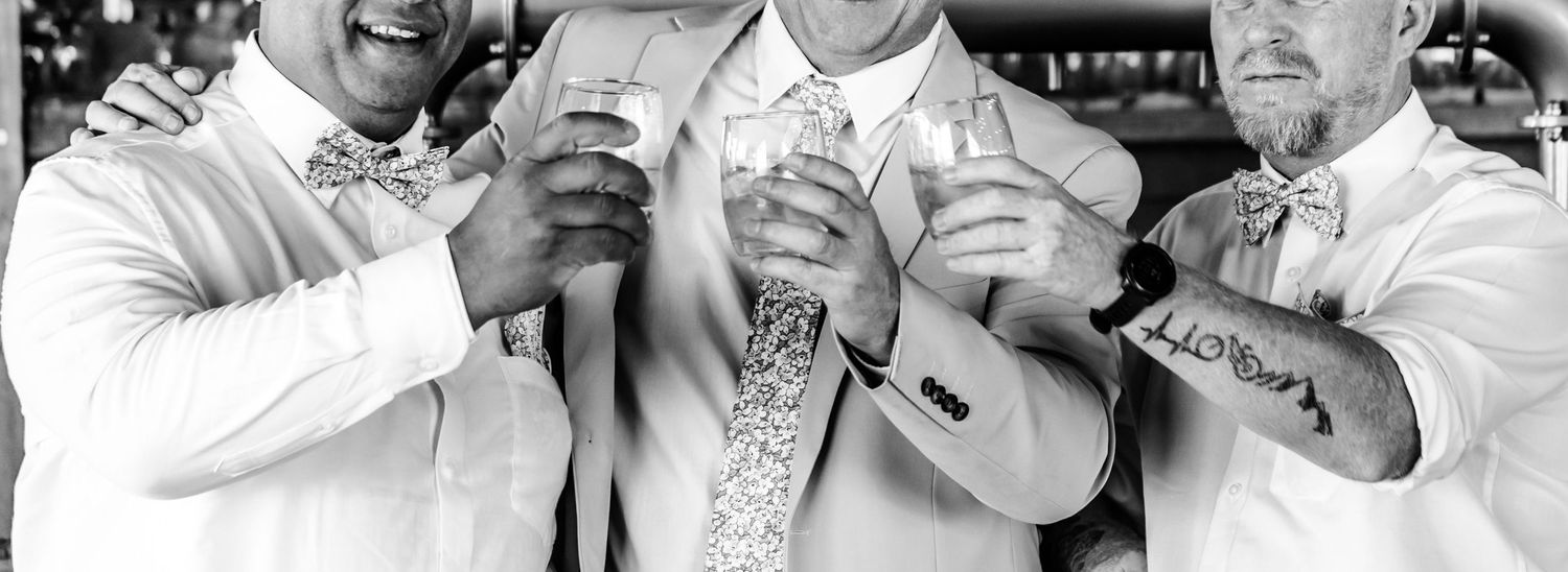 Three men in casual attire hold up their drinks while smiling in a black and white candid photograph.
