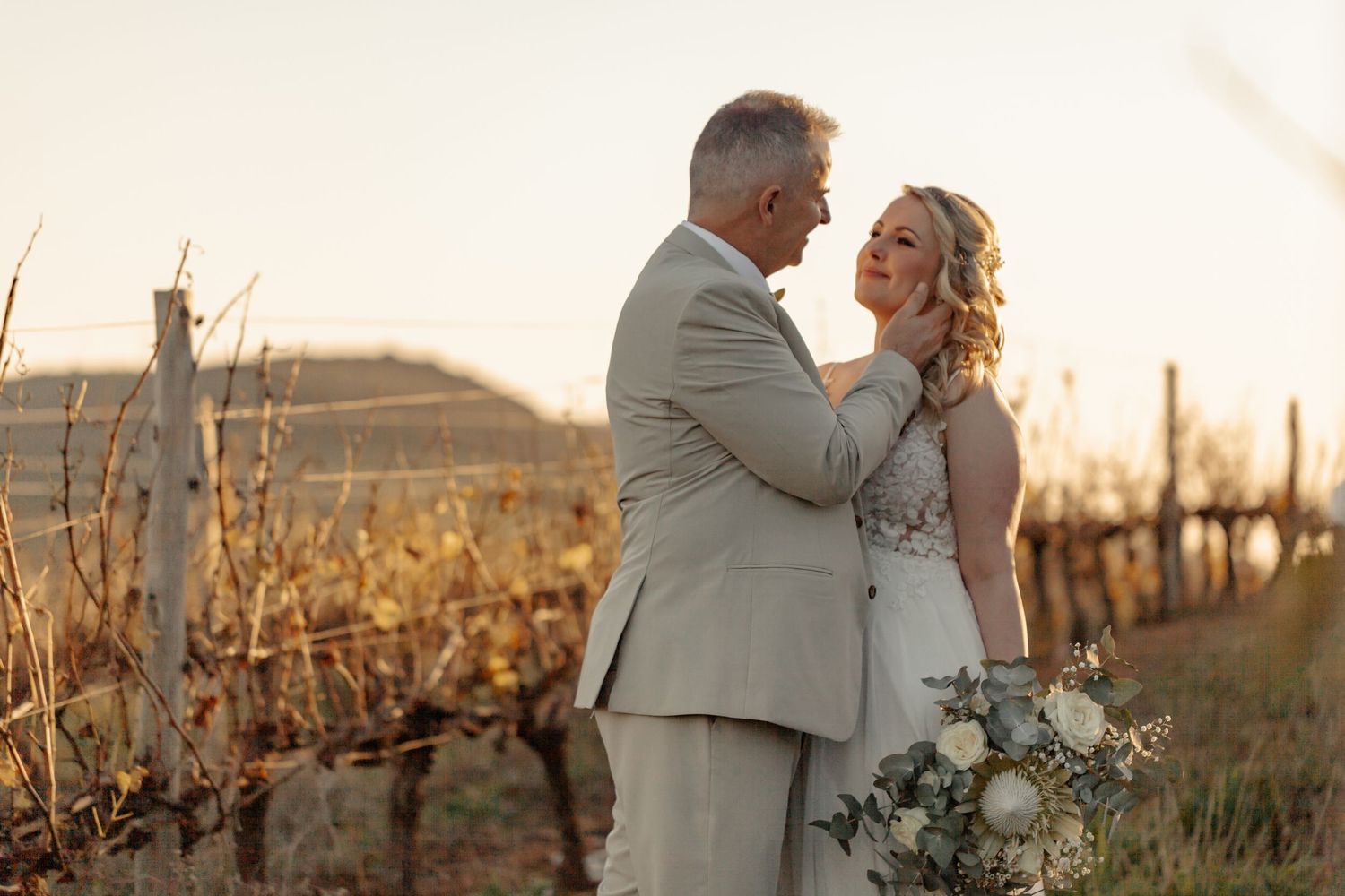 A couple shares a romantic embrace at sunset in a vineyard with golden sunlight illuminating the scene.