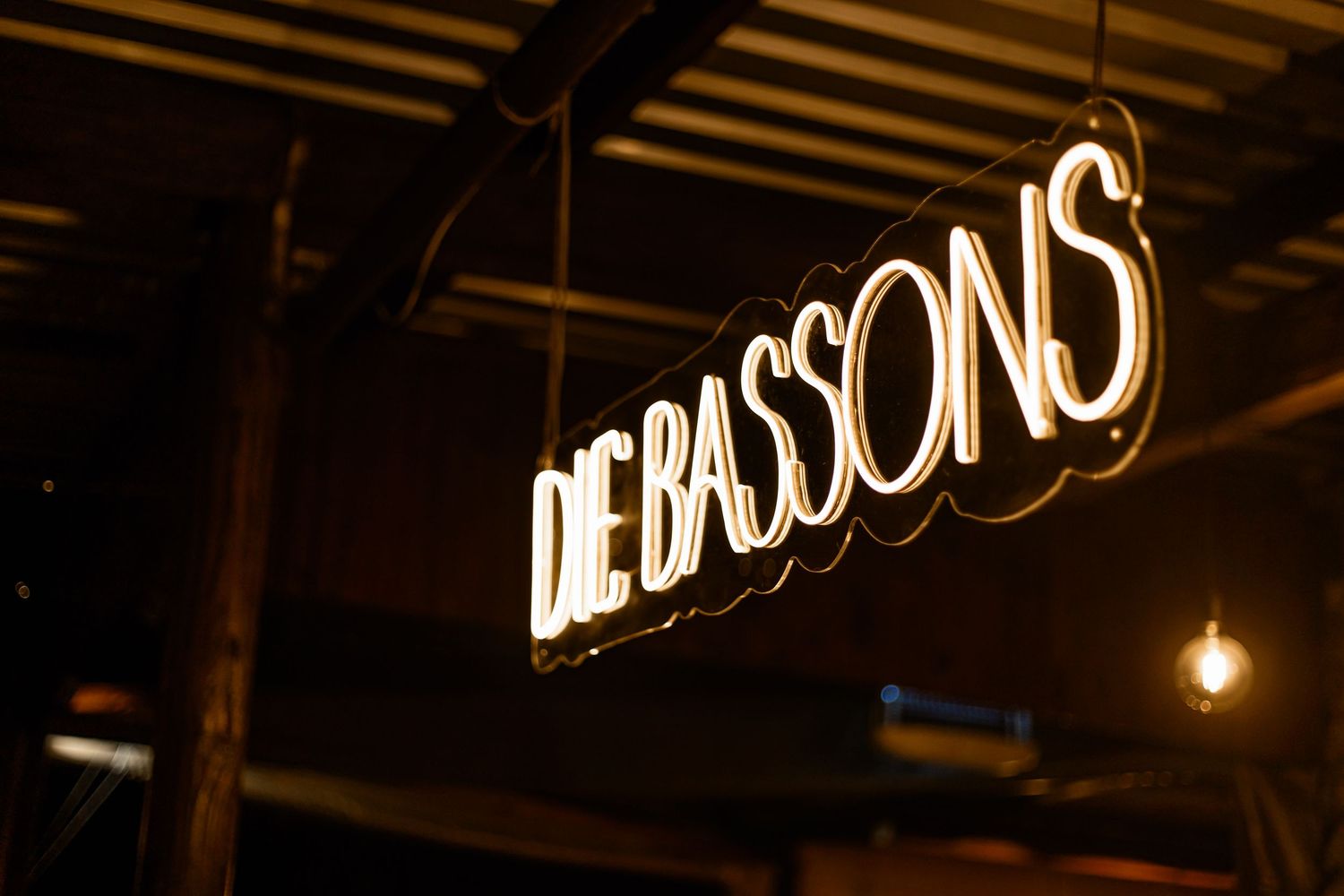 Illuminated 'Le Bassons' restaurant sign glows in white neon letters against a dark ceiling with pendant lighting.