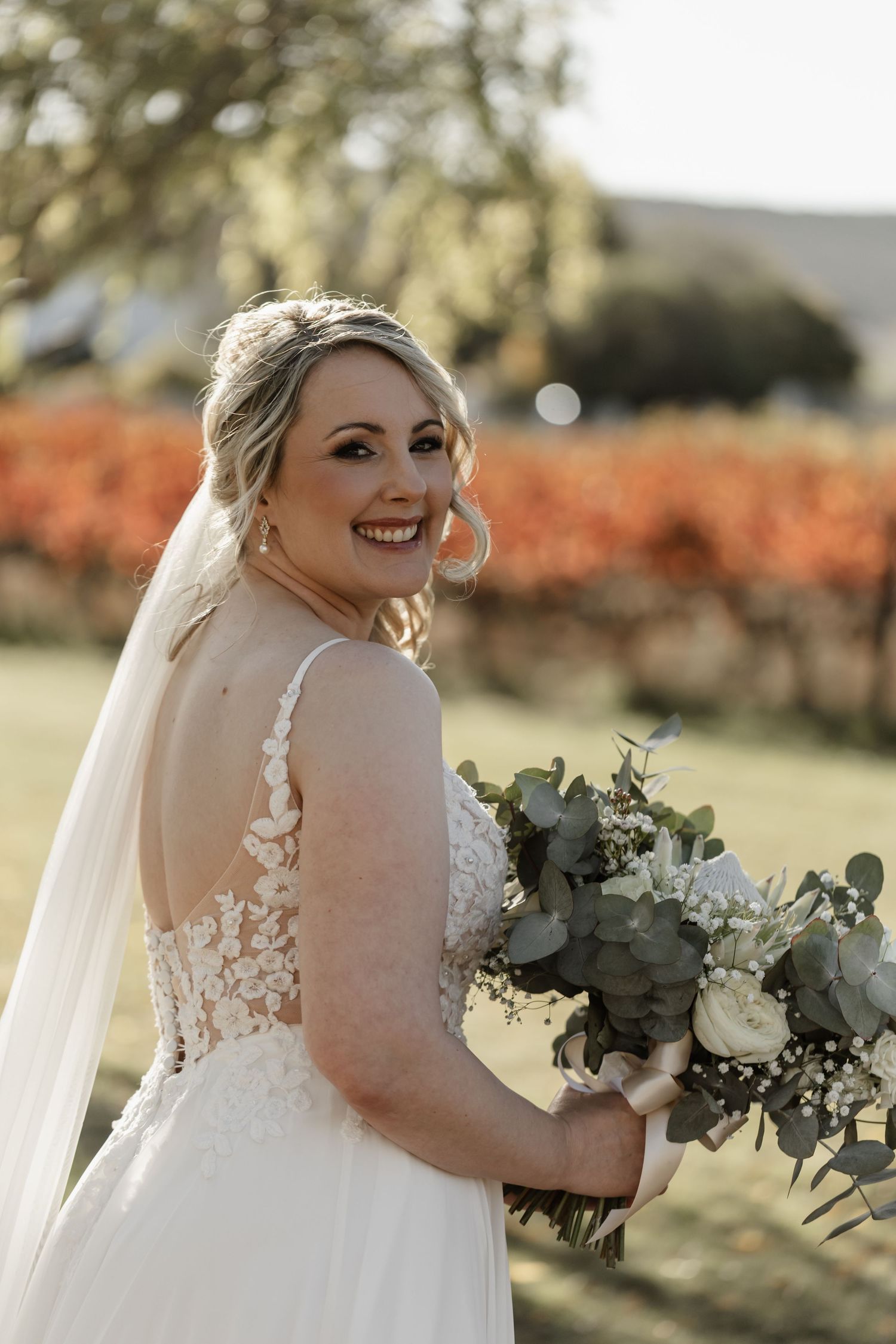 Beautiful bride wearing lace wedding dress poses with eucalyptus and white rose bouquet against autumn vineyard backdrop.
