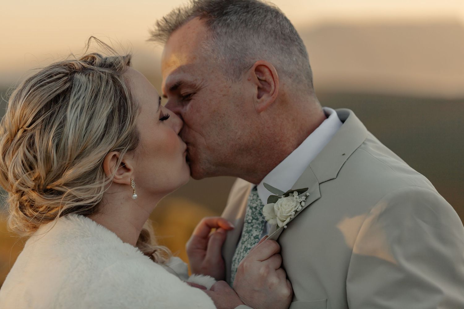Romantic couple embrace and kiss during sunset at their outdoor wedding with beautiful golden lighting.