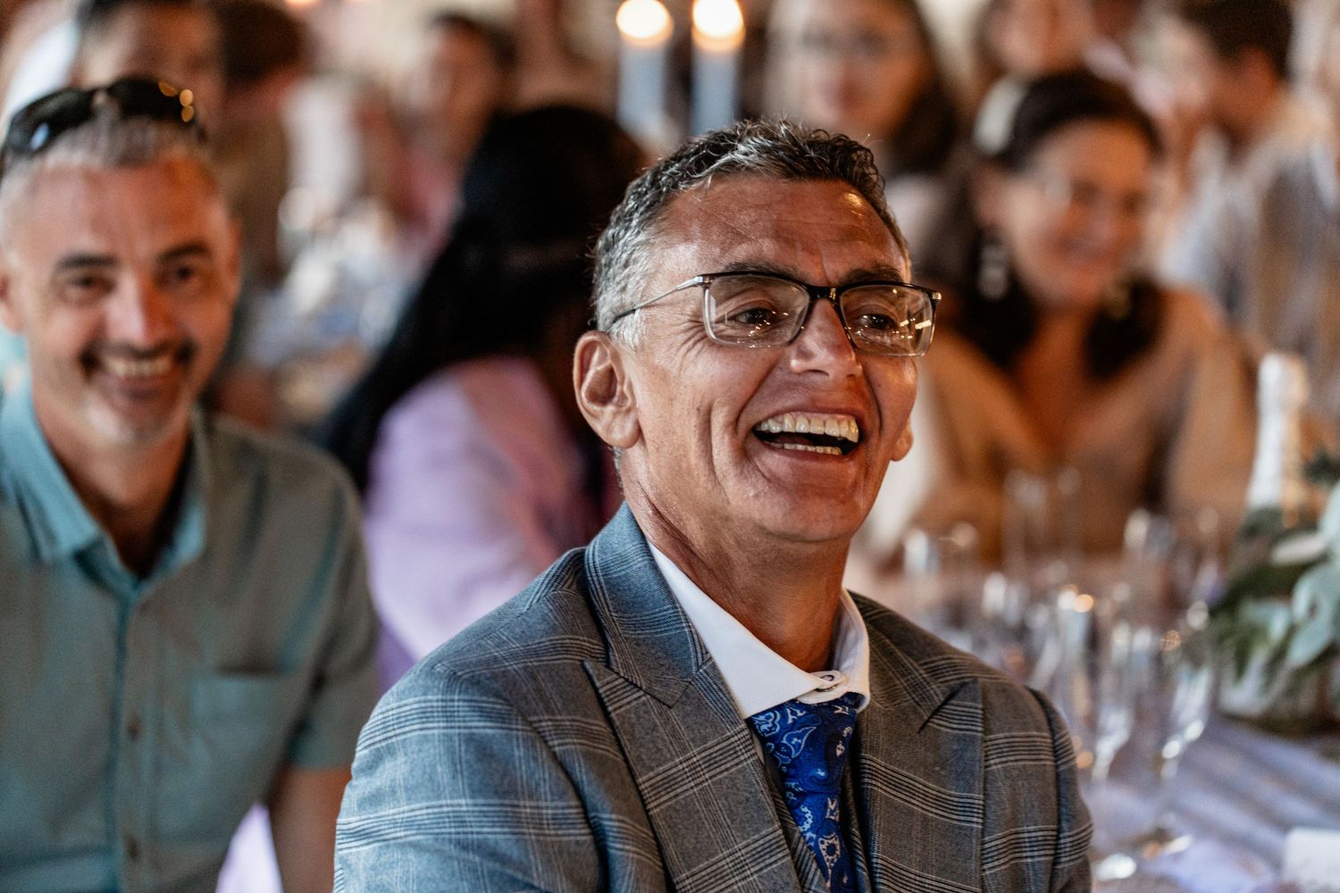 Laughing wedding guest in grey suit and blue tie sitting at reception dinner table among other attendees.