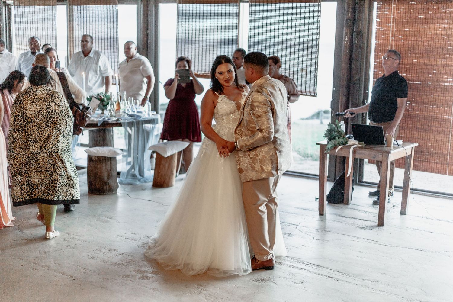 A couple shares their first dance at a rustic industrial wedding venue with guests watching from the sidelines.