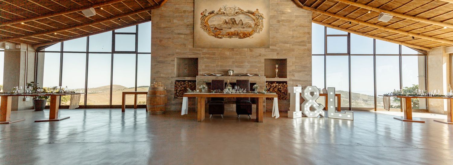 Modern rustic event venue interior with high wood beam ceiling, stone fireplace, and panoramic windows overlooking scenic views.