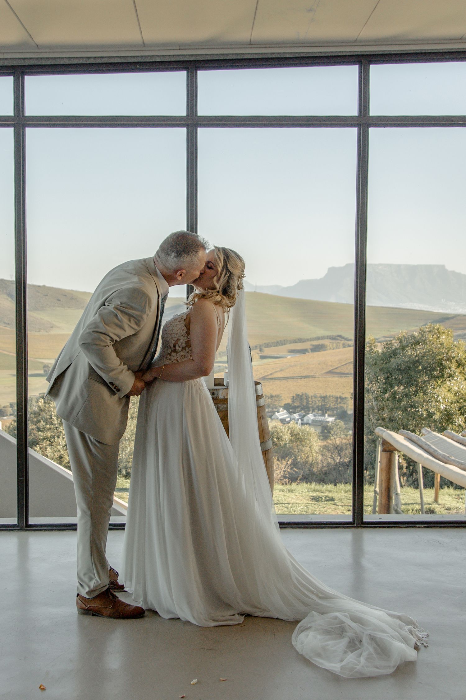 Romantic wedding couple shares a dip-style kiss in front of large windows overlooking mountainous landscape.