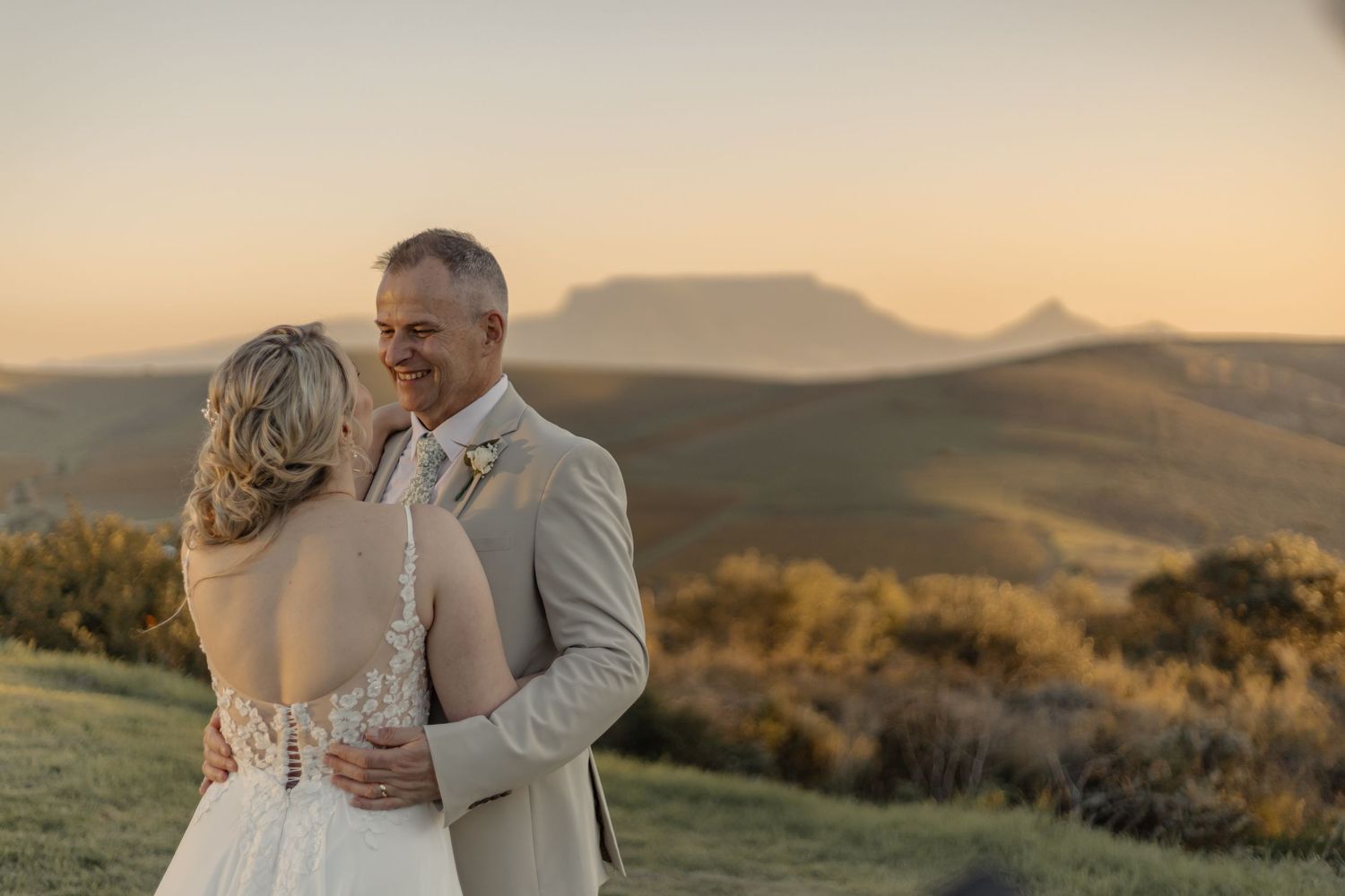 Newlyweds embrace during golden hour in scenic mountain landscape at sunset while wearing formal wedding attire.