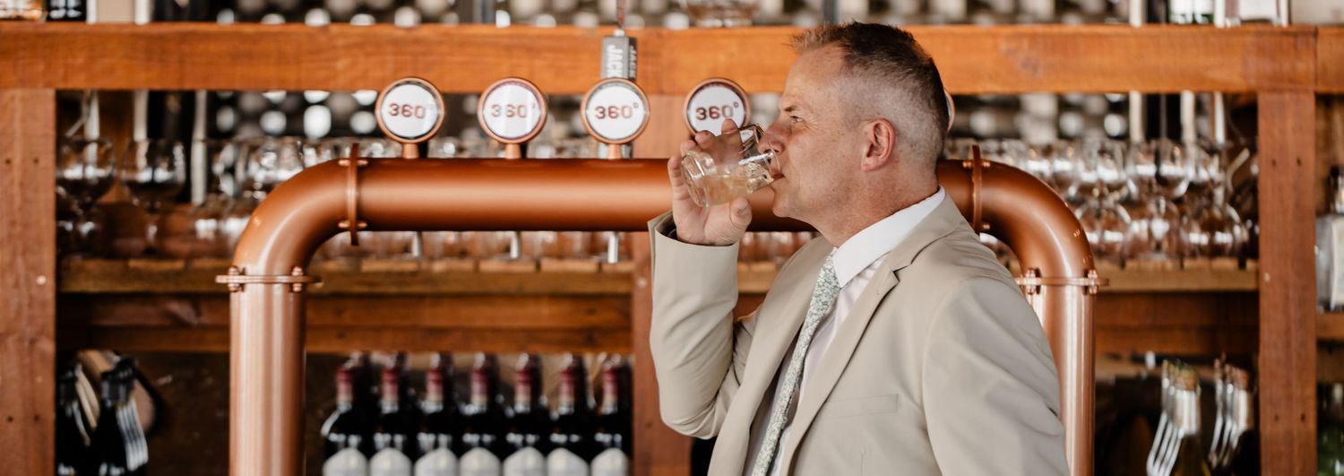 A person in a light suit sits at a wooden bar counter and sips from a copper mug near bottles and glassware.