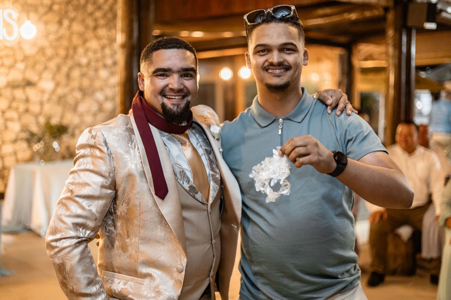 Two friends pose together at an indoor event, one wearing a metallic blazer and the other in a gray t-shirt.