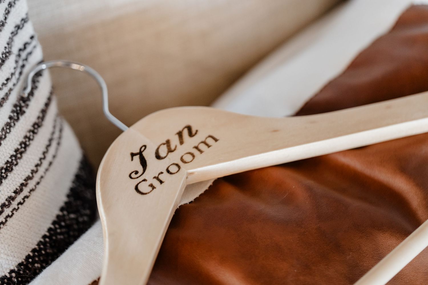 A wooden wedding garment hanger with 'Fan Groom' text engraved on light wood against brown leather and fabric.