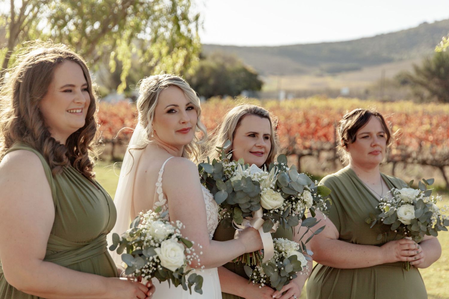 Four bridesmaids in olive green and white dresses hold bouquets while standing in front of autumn vineyard at sunset.