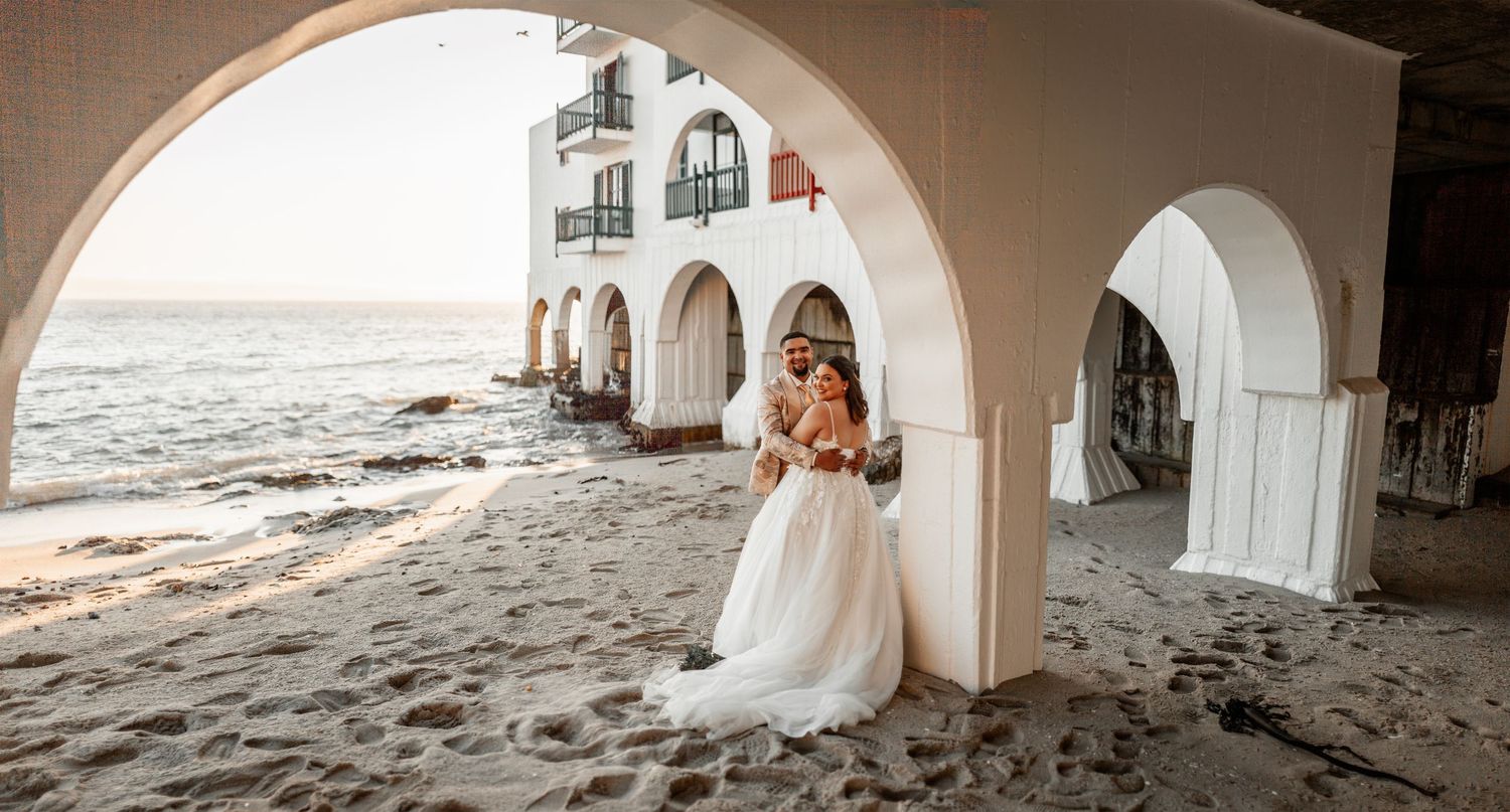 A romantic coastal view through white Mediterranean-style arches with ocean waves crashing against the sandy beach.