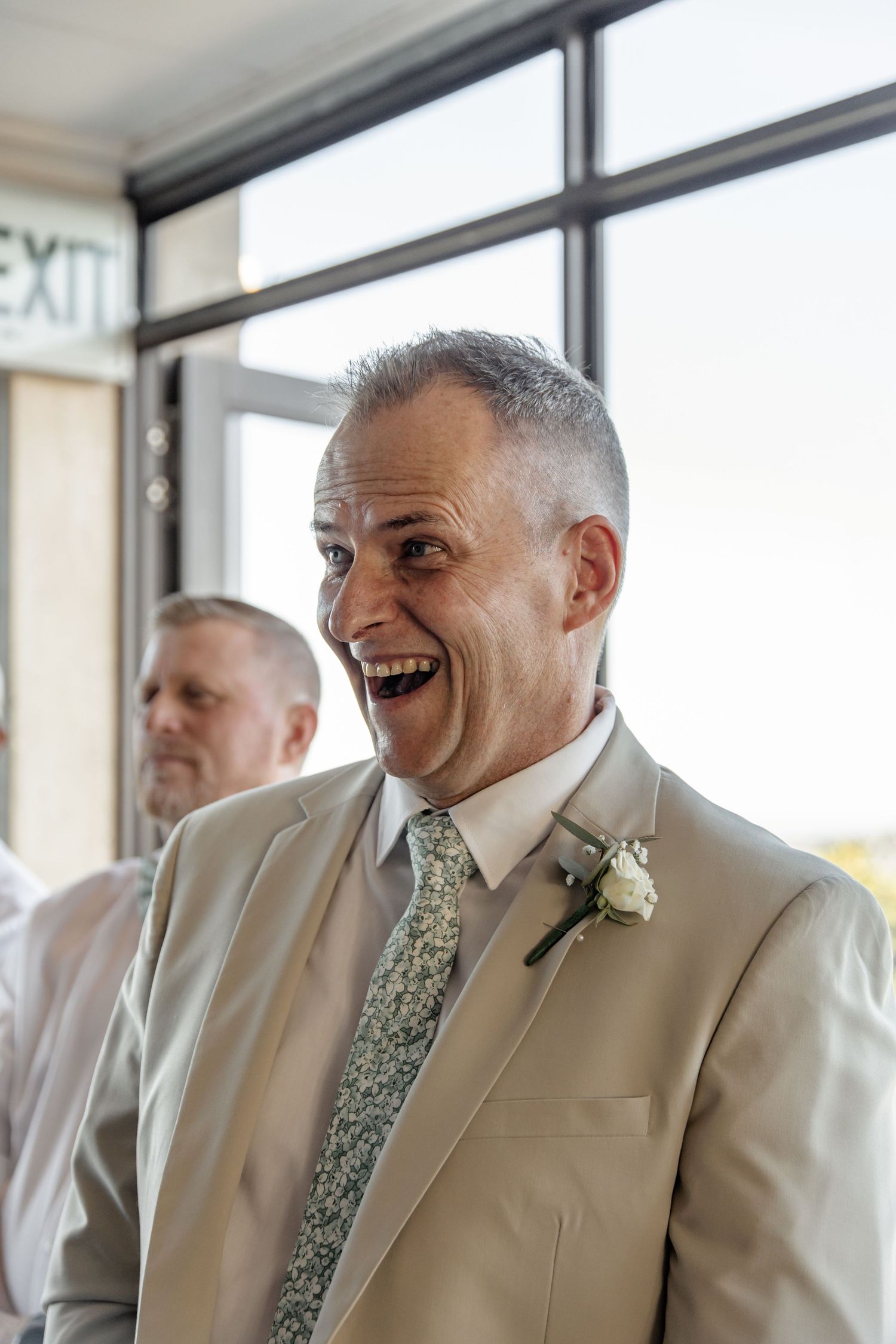 A guest at a wedding ceremony wearing a tan suit and floral tie laughs joyfully near an exit sign.