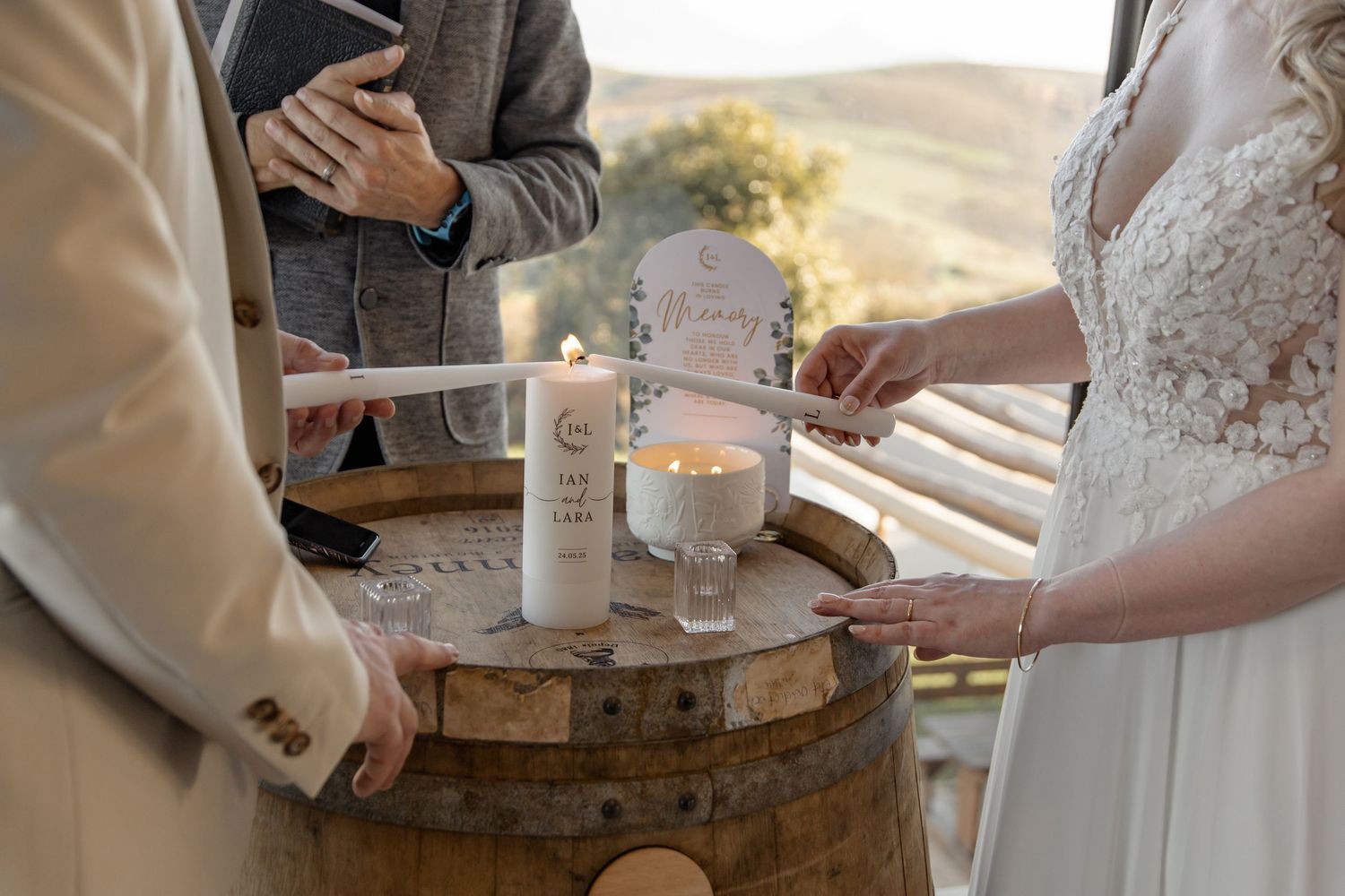 Couple performs unity candle ceremony with white candles on rustic wooden barrel during outdoor wedding ceremony.