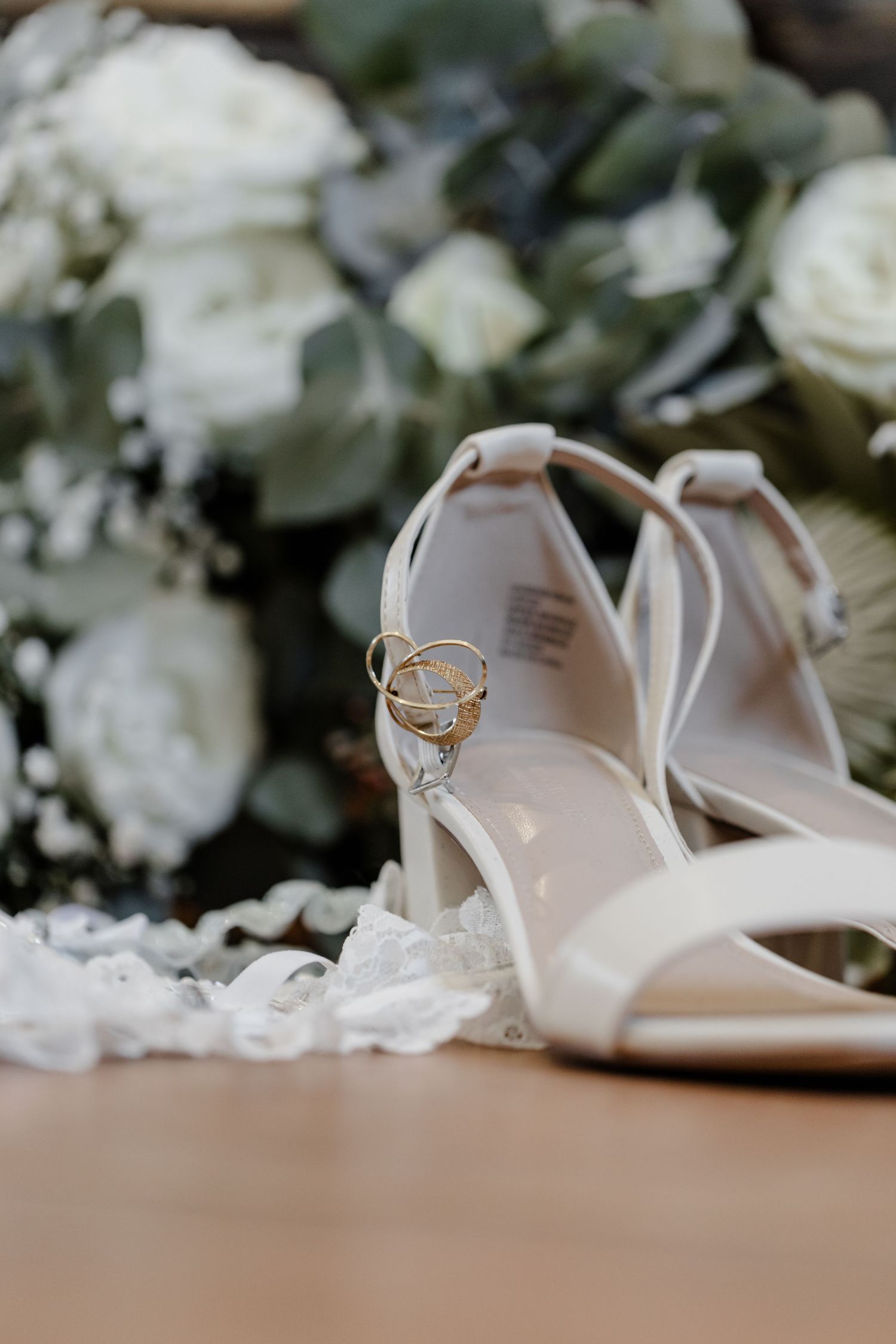 Elegant white bridal sandals with wedding rings displayed among white roses and eucalyptus leaves.