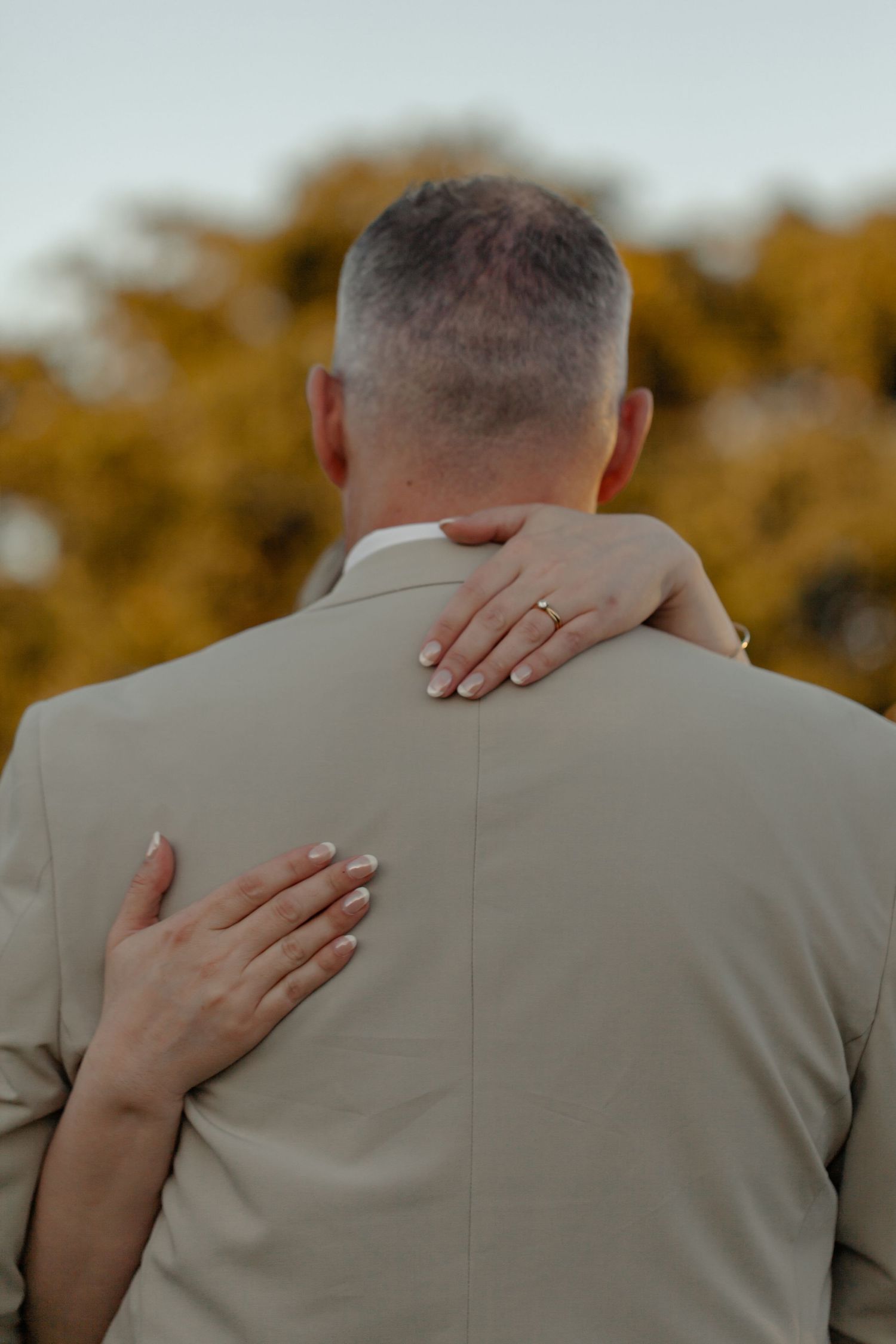 Hands with manicured nails embrace the back of someone wearing a beige suit jacket against an autumn backdrop.