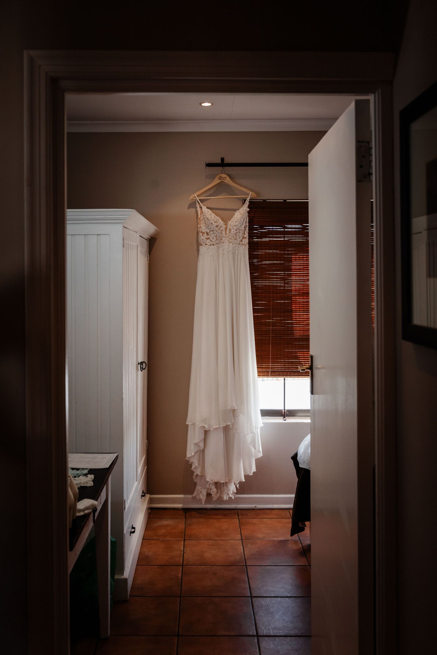 A white wedding dress hangs elegantly in a doorway against a wooden floor and dark neutral walls.