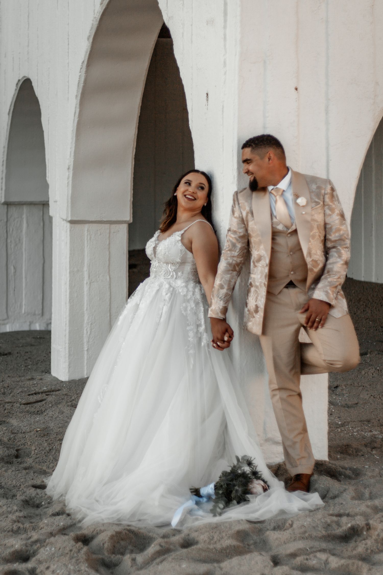 A couple shares a joyful moment on a sandy beach with white archways, wearing a wedding dress and tan suit.