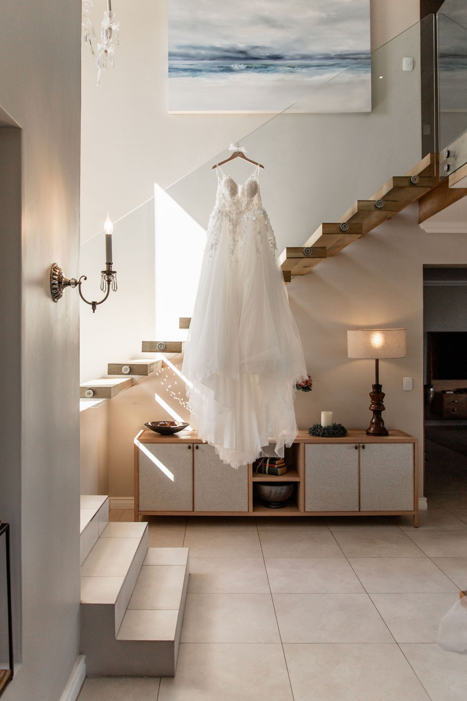 Elegant white wedding dress hanging in modern minimalist entryway with wooden stairs and coastal artwork.
