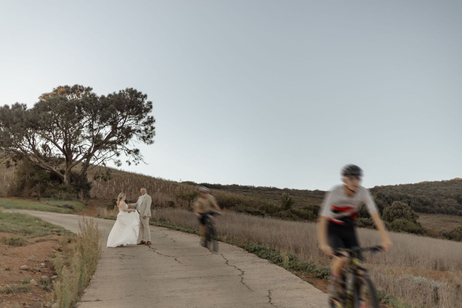 A person riding a bike on a dirt path while figures in motion walk in the opposite direction near a tree at sunset.