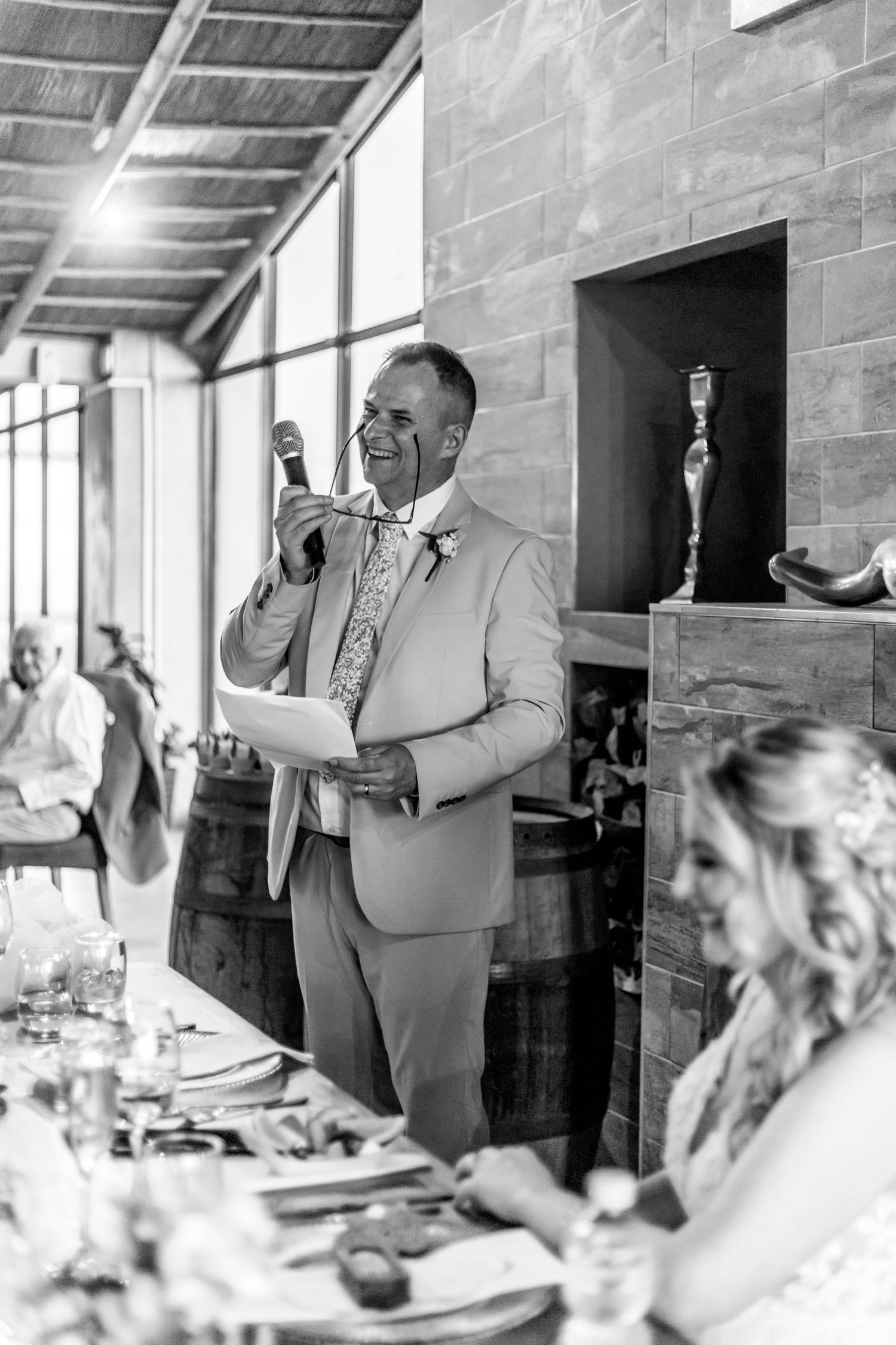 Black and white photo of a wedding reception speech with speaker in light suit at dining table indoors.
