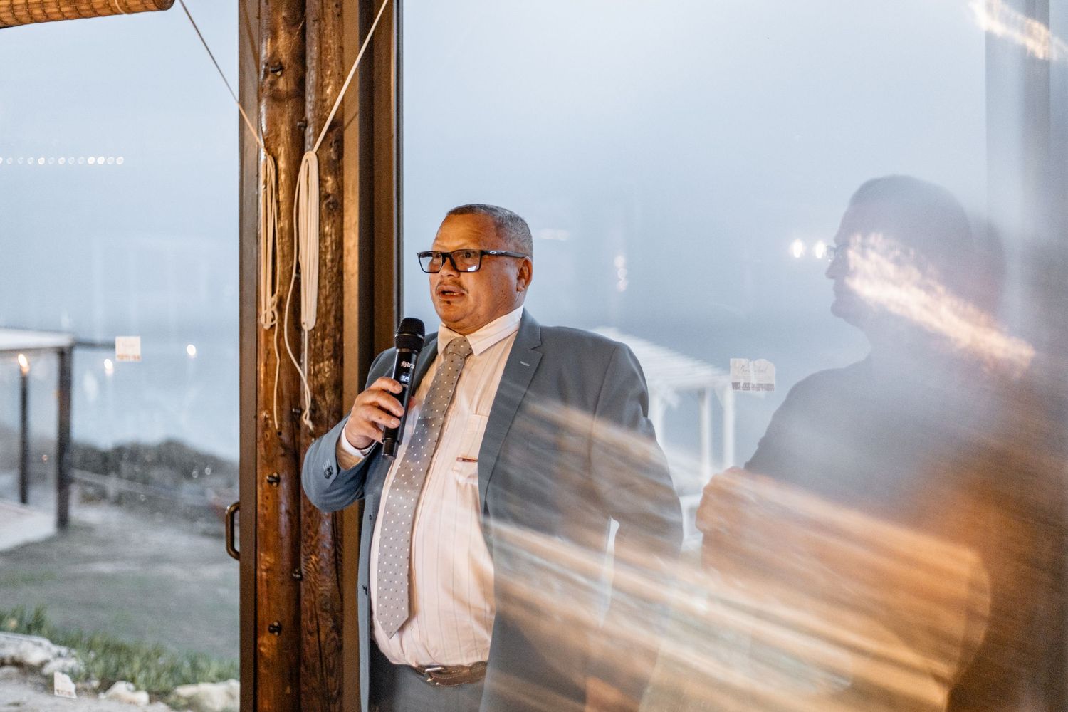 Someone in glasses and a suit jacket holds a microphone while speaking at an evening event venue with rustic wooden beams.