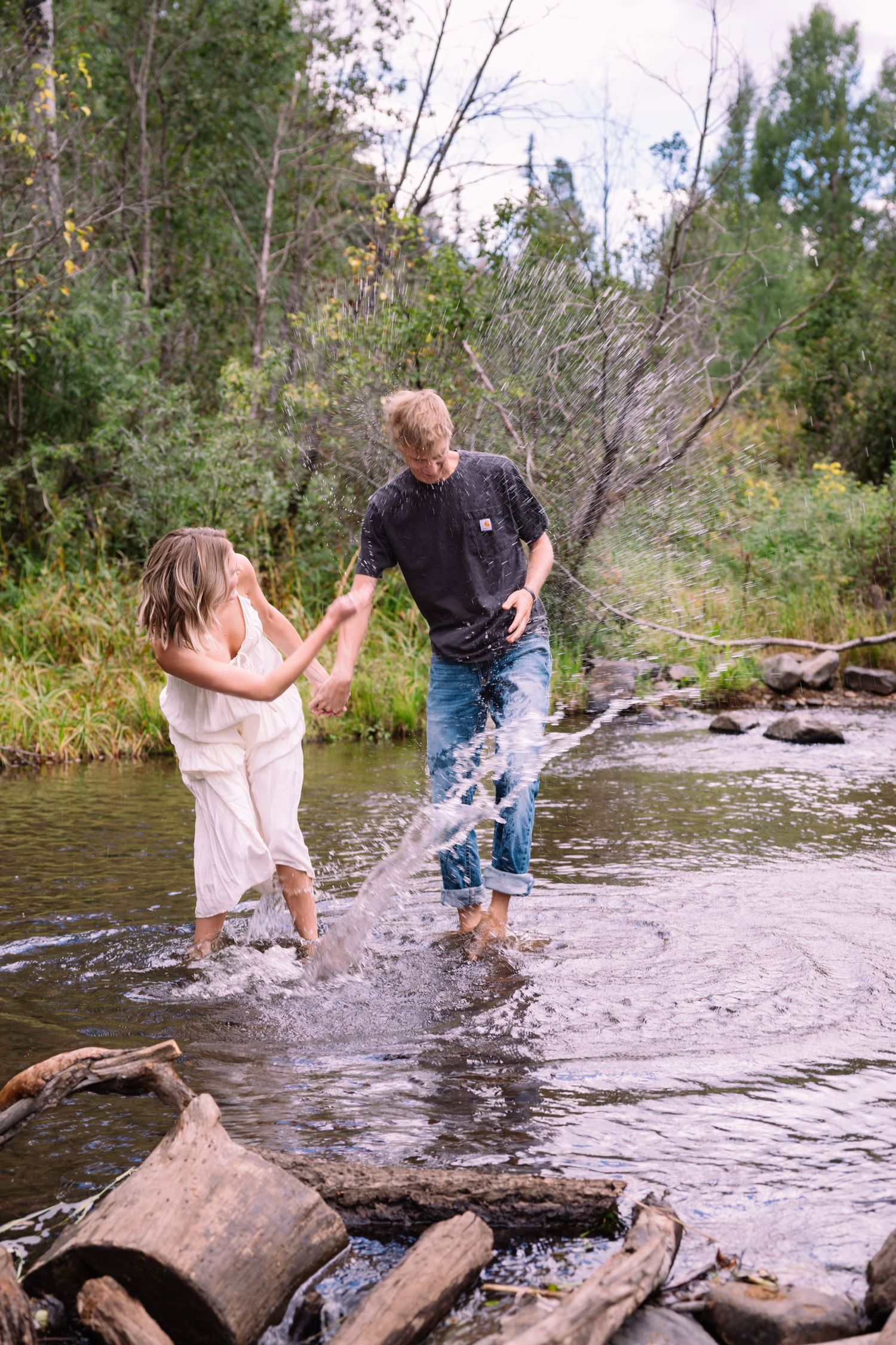 Two people splash and play in a shallow stream on a sunny day, surrounded by rocks and trees in a natural setting.