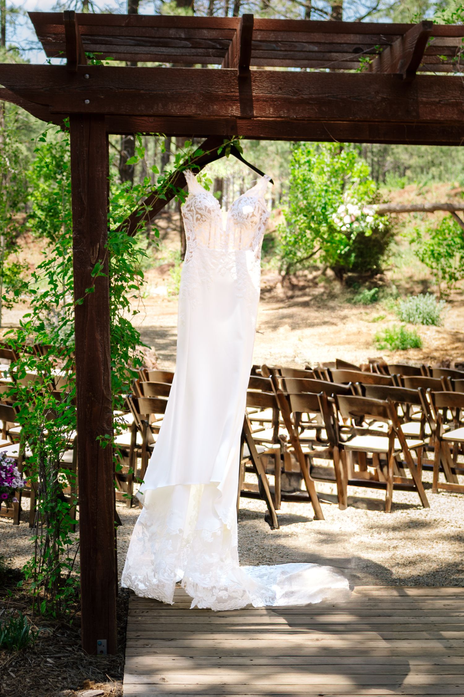 A white lace wedding dress hangs from a wooden pergola in an outdoor garden ceremony setting with folding chairs.