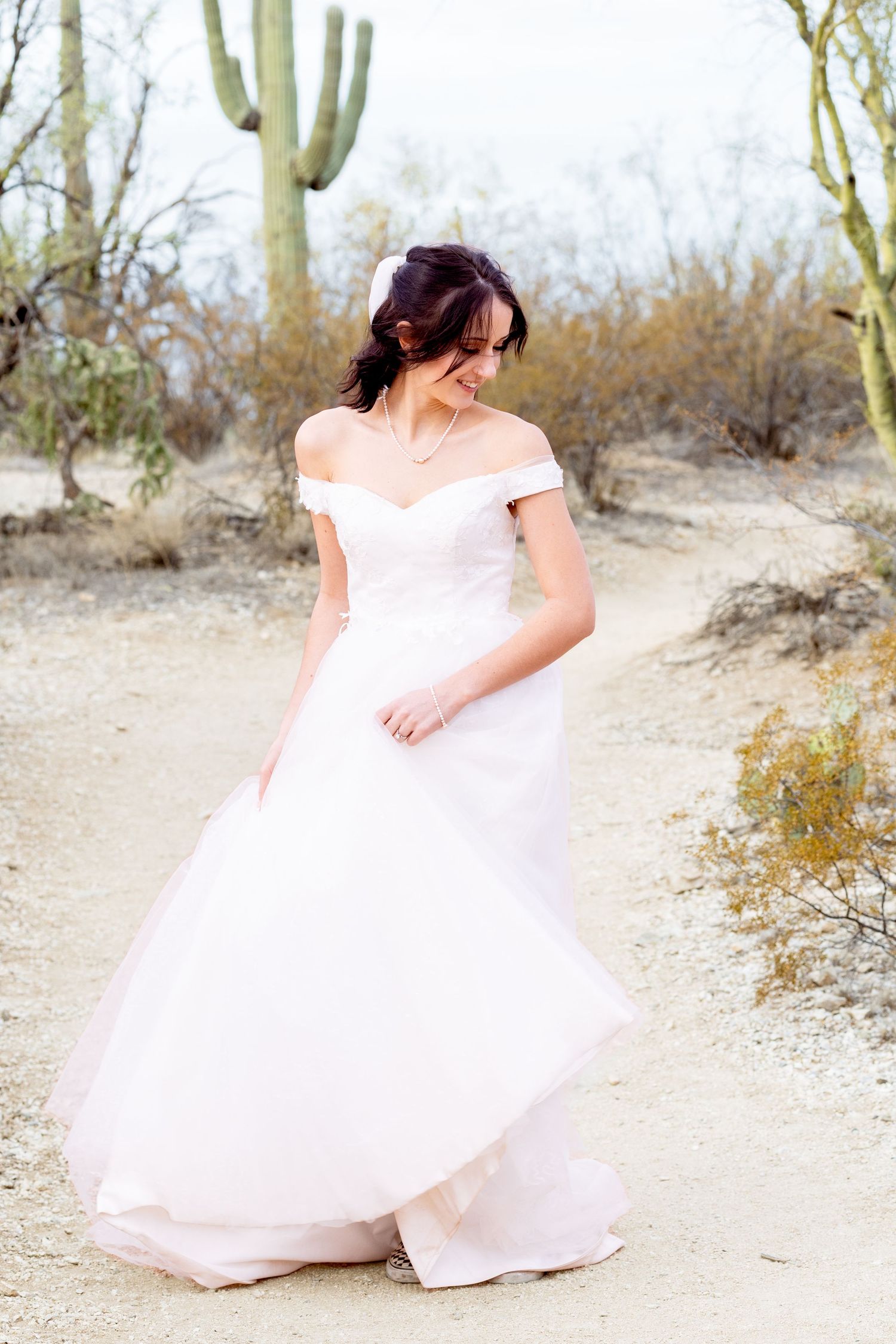 Bride in flowing white dress twirling on desert path surrounded by cacti.