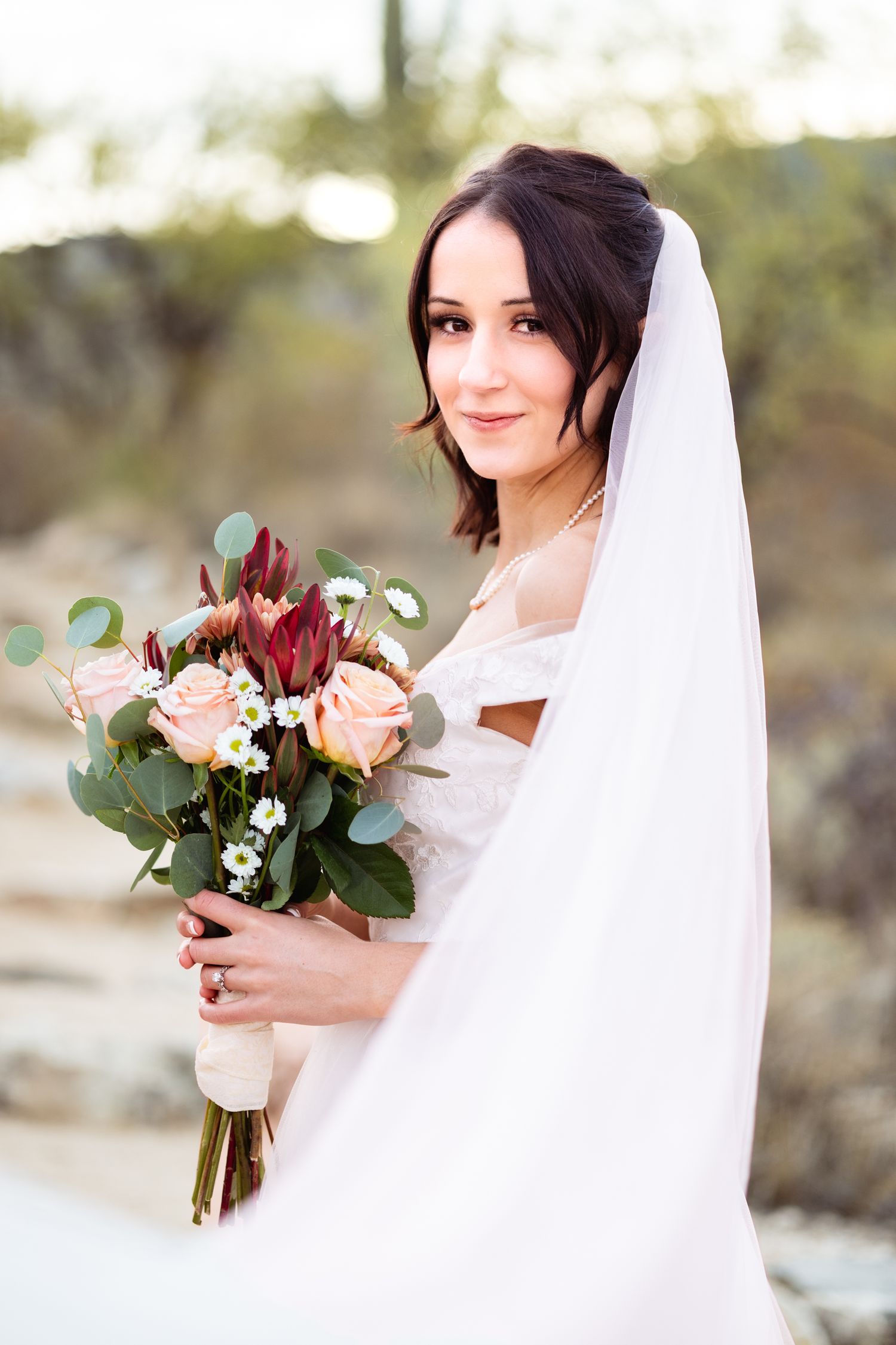 Bride holding romantic bouquet of peach roses and burgundy flowers against desert landscape.