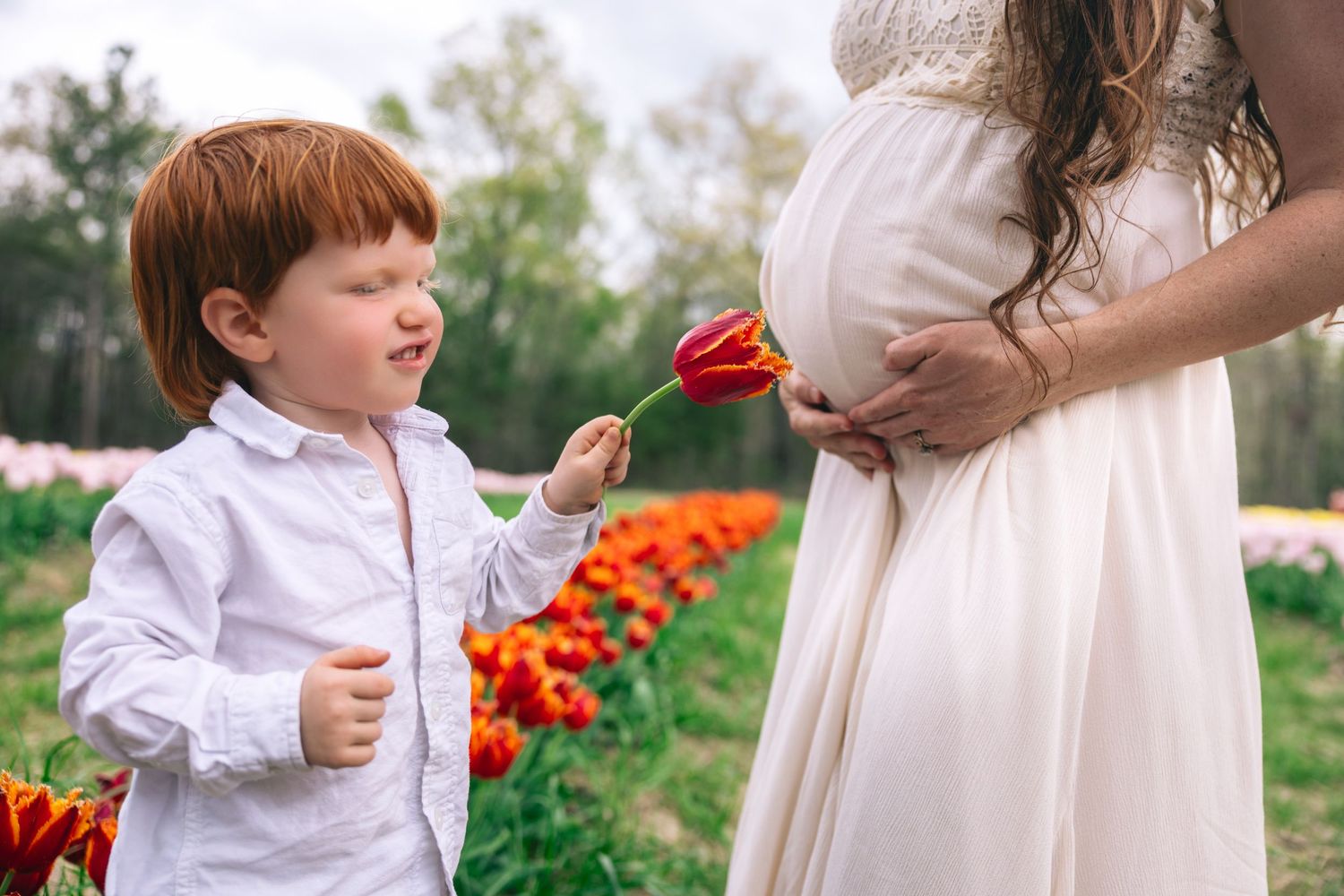 Child in white dress smelling flowers in garden while standing next to adult holding bouquet of orange marigolds.
