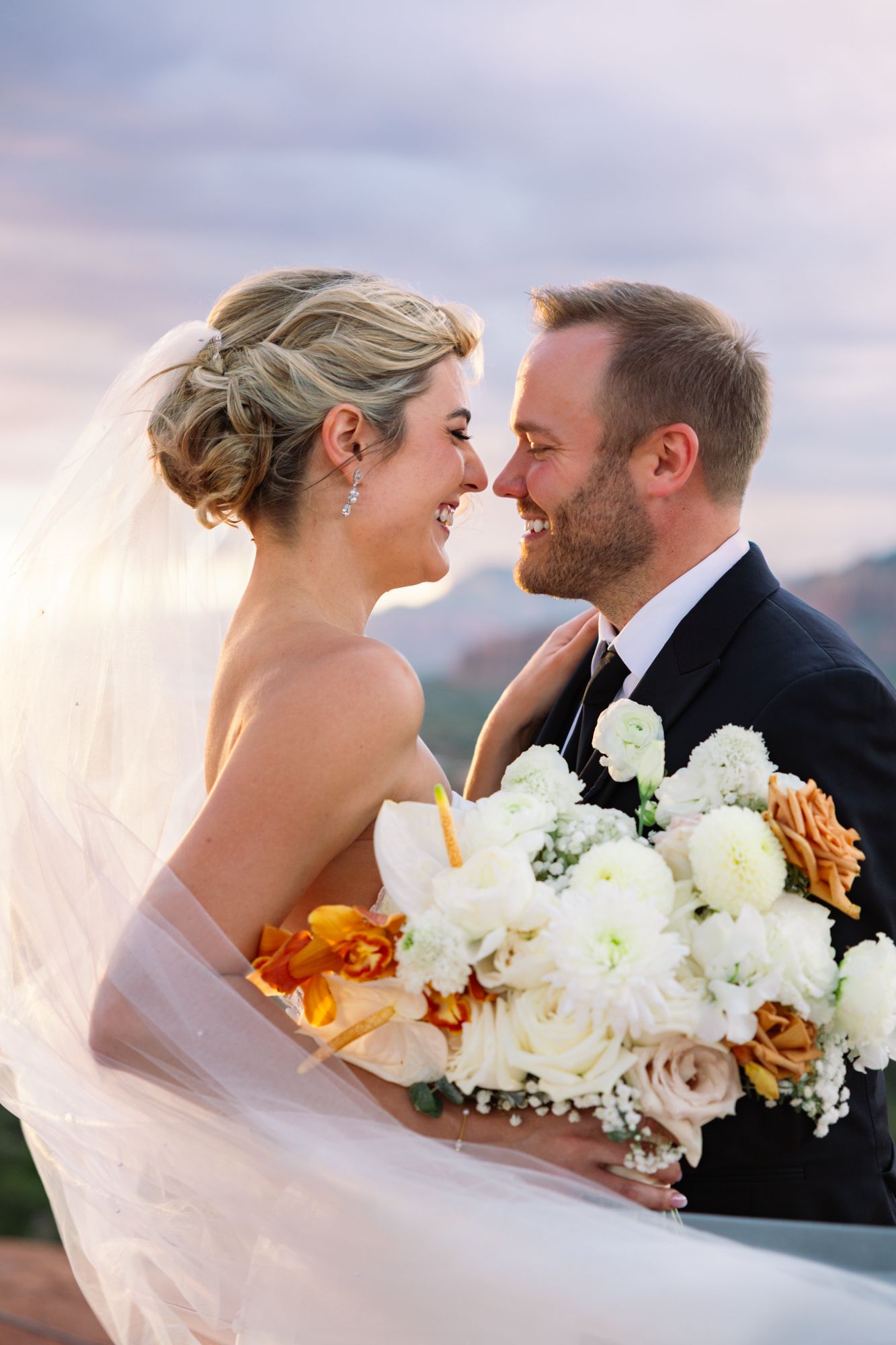 A romantic wedding moment with white and orange bouquet captured at sunset in front of a mountain landscape.