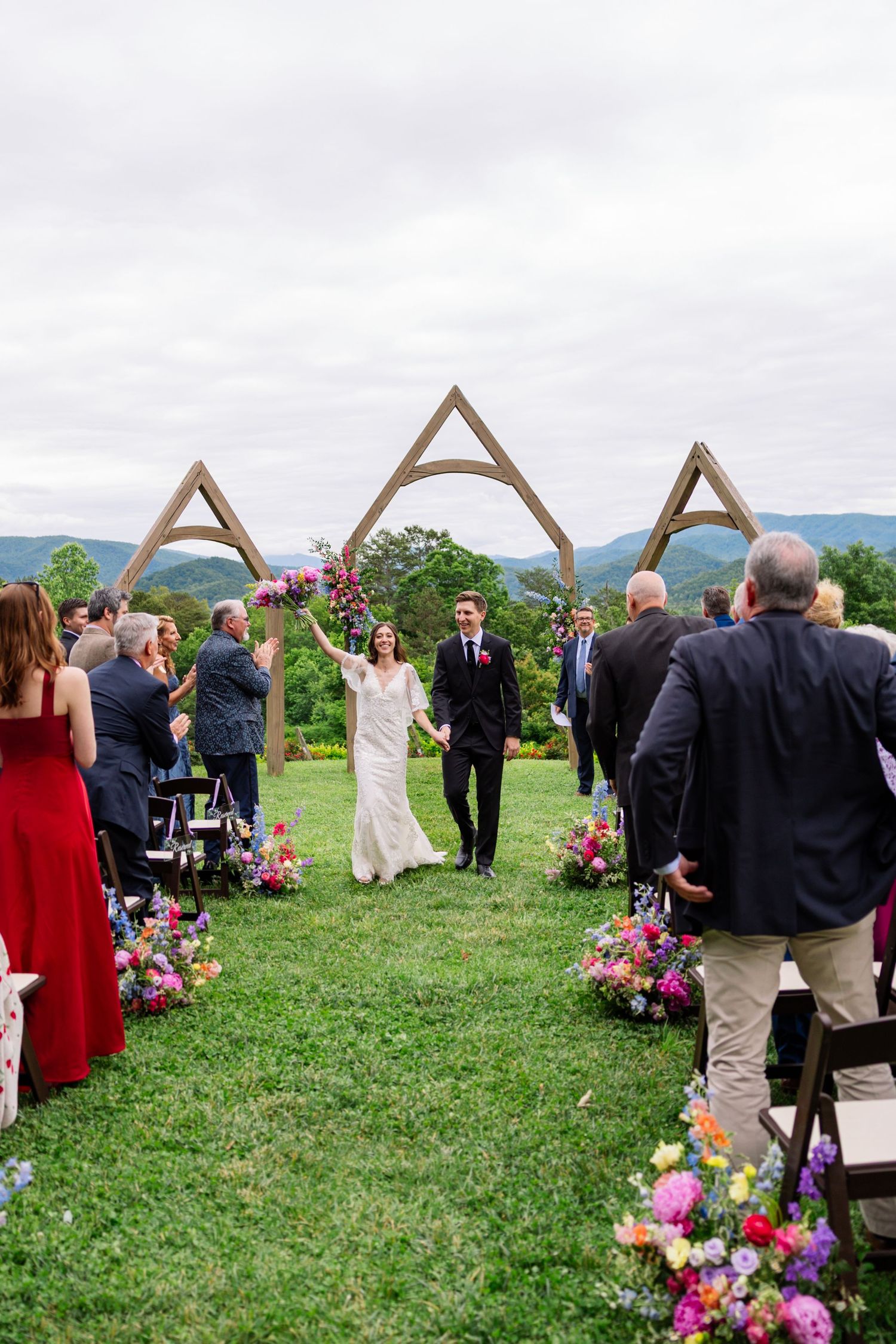 Two people walk down an outdoor aisle lined with colorful flowers under triangular wooden arches in a mountain setting.