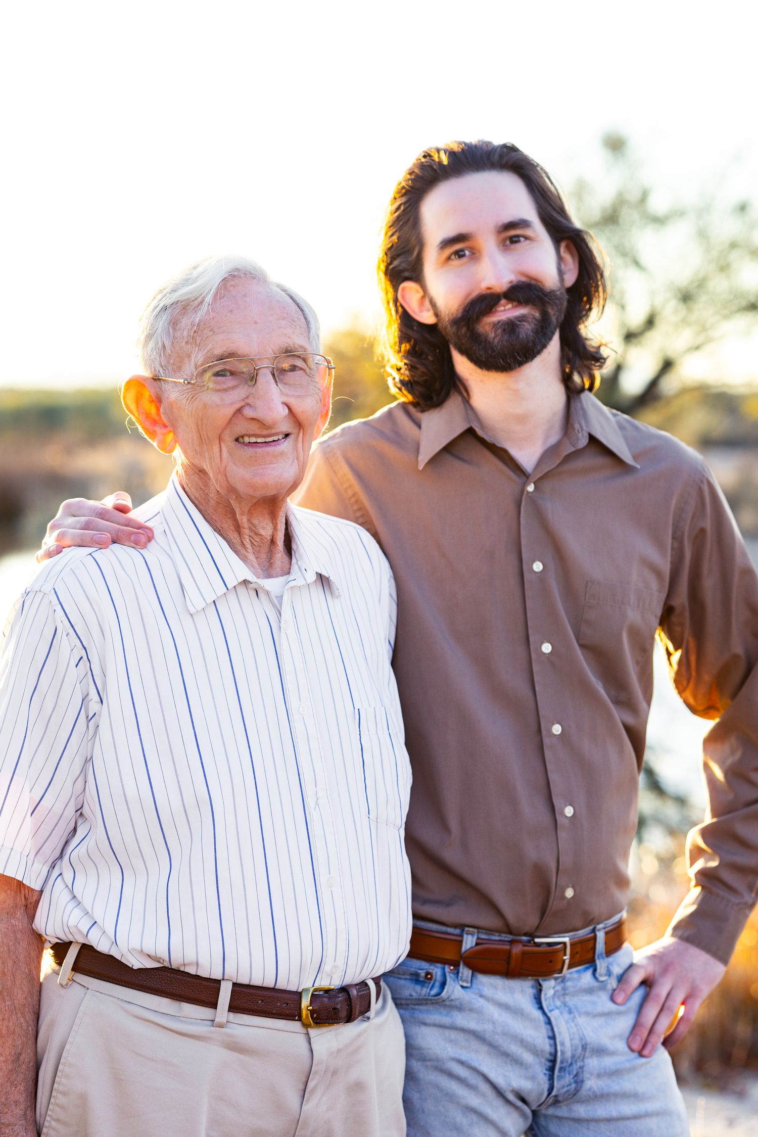 Two individuals photographed outdoors during golden hour, one elderly wearing stripes and one younger in brown.