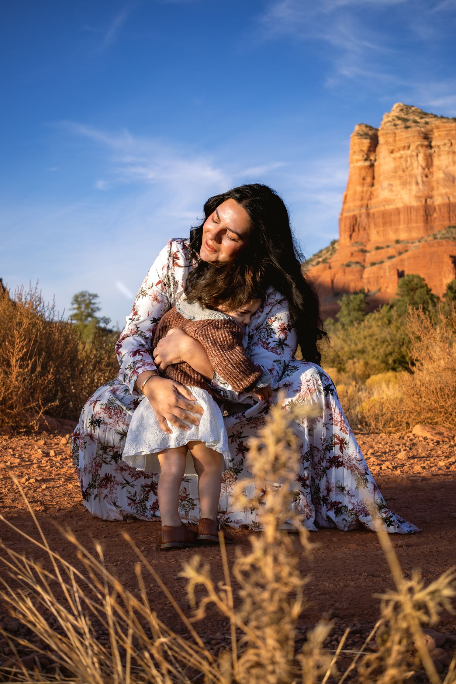 Woman in floral dress sits on red soil with towering sandstone cliffs in background.
