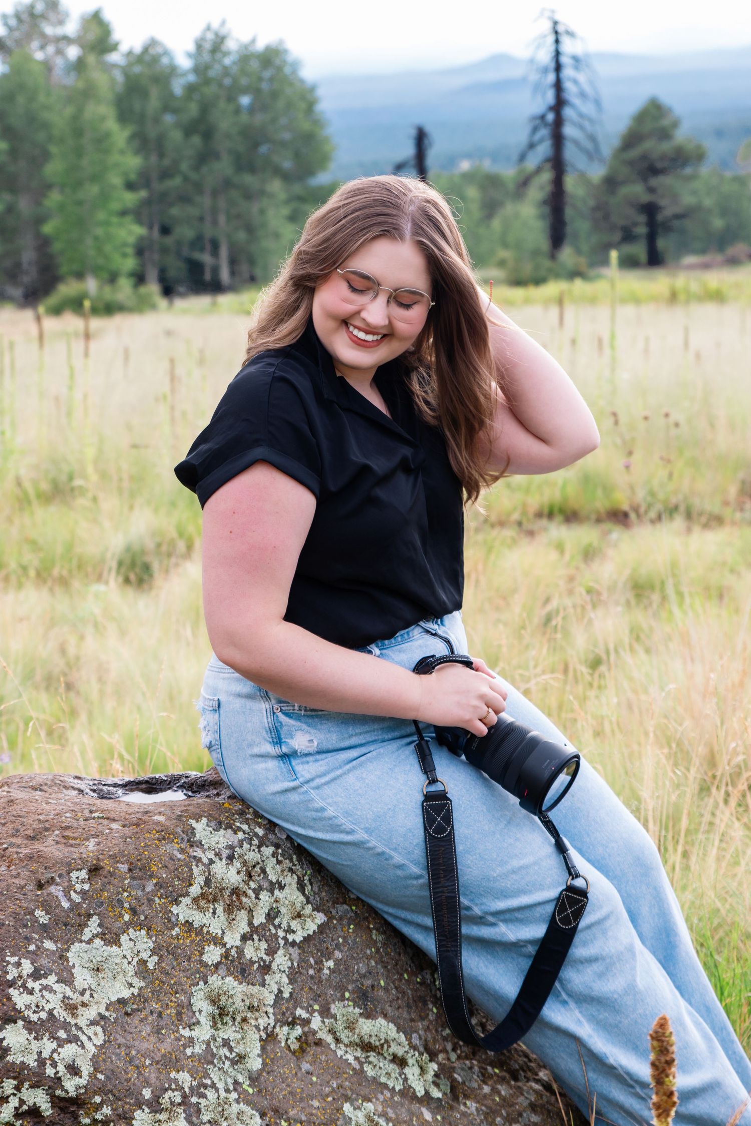 Bethany, sitting in a black shirt and jeans, sits casually on a large rock in a field. She is a Knoxville photographer