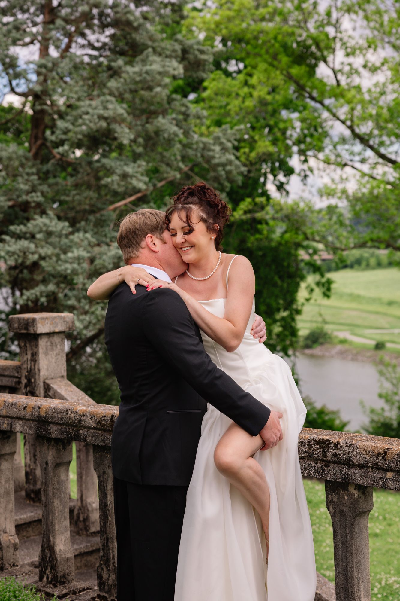 A romantic wedding couple embraces on a stone balcony surrounded by lush green trees and gardens.