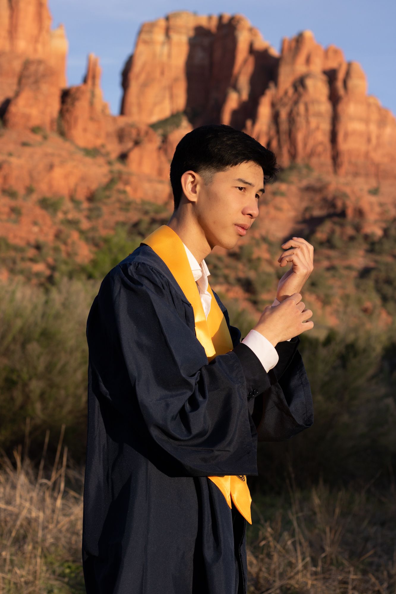 Graduate in navy blue gown and yellow stole poses in front of red rock formations.