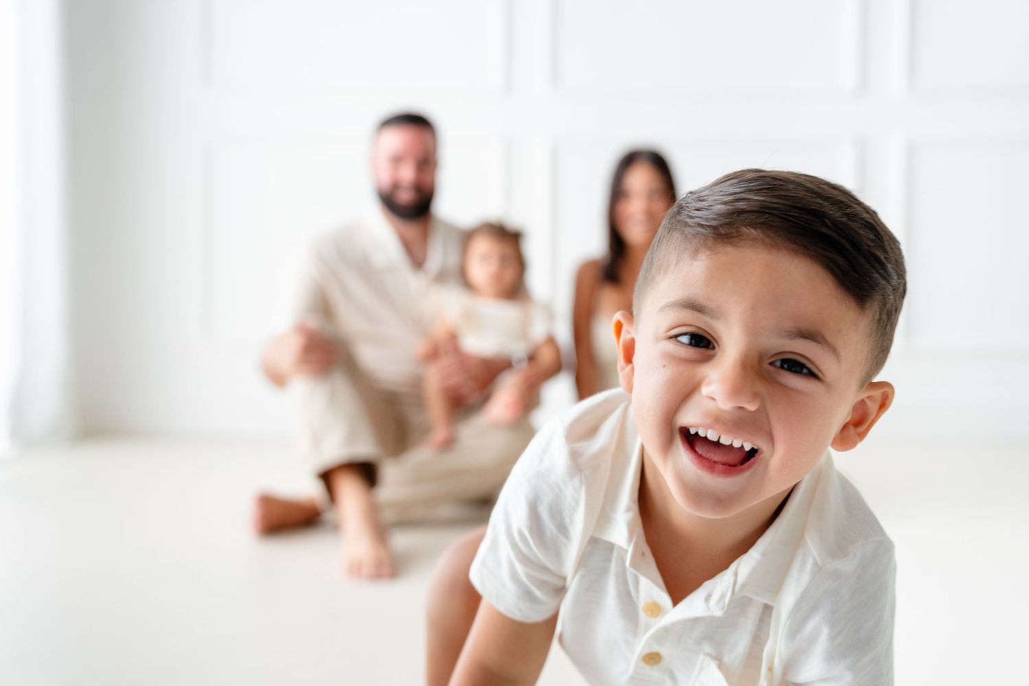 A joyful child in a white shirt laughs while family members sit in the background of a bright white room.