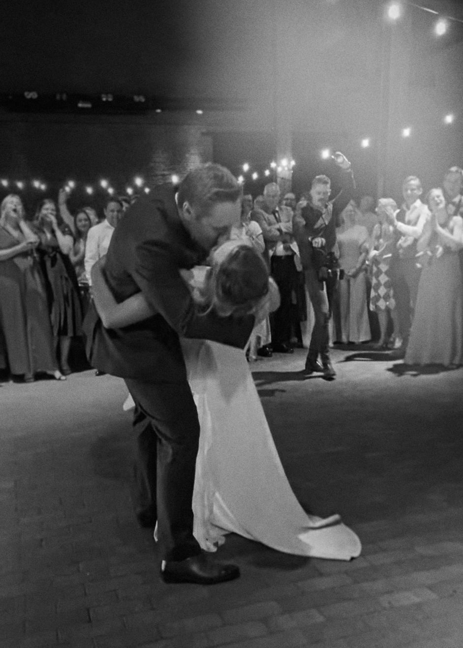 A couple shares a romantic dip during their first dance at a wedding reception with string lights and guests looking on.
