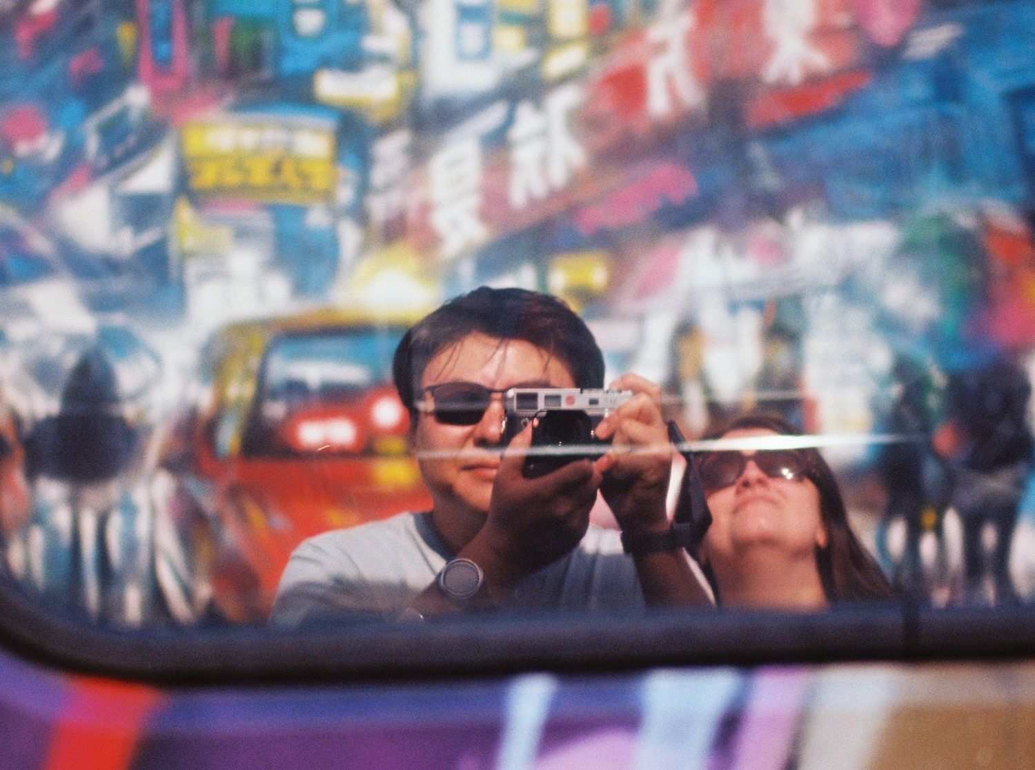 Self portrait reflection in car mirror with street art and neon signs in background.