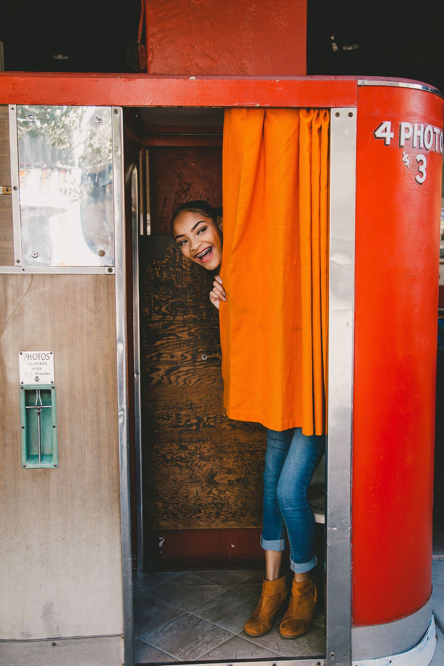 Someone peeks out playfully from behind an orange curtain in a vintage photo booth wearing jeans and brown boots.