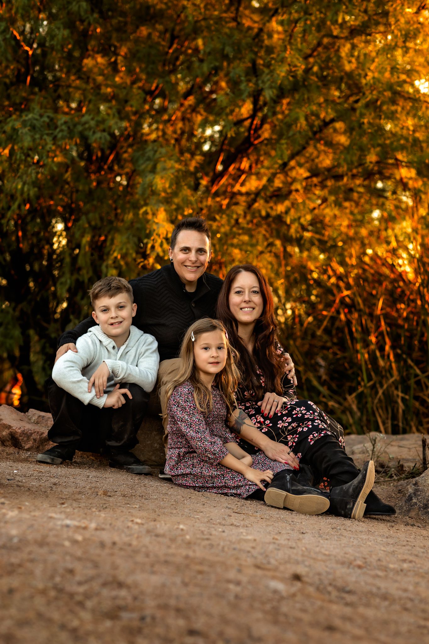 A family sits together on rocks during autumn with colorful fall foliage glowing warmly in the background for their fall photo session.