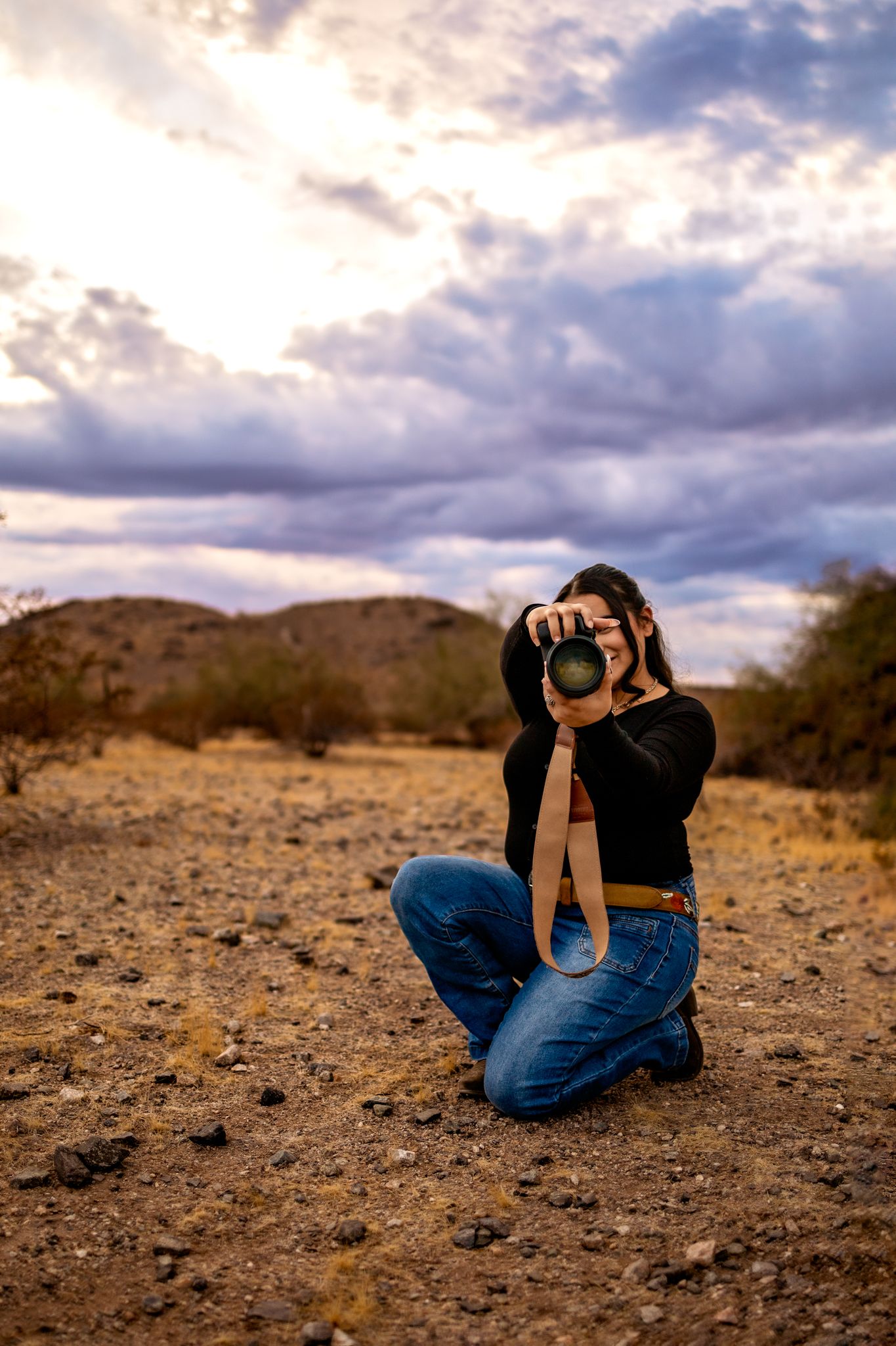 A photographer in black clothing takes photos in a desert landscape during sunset holding her Canon camera.