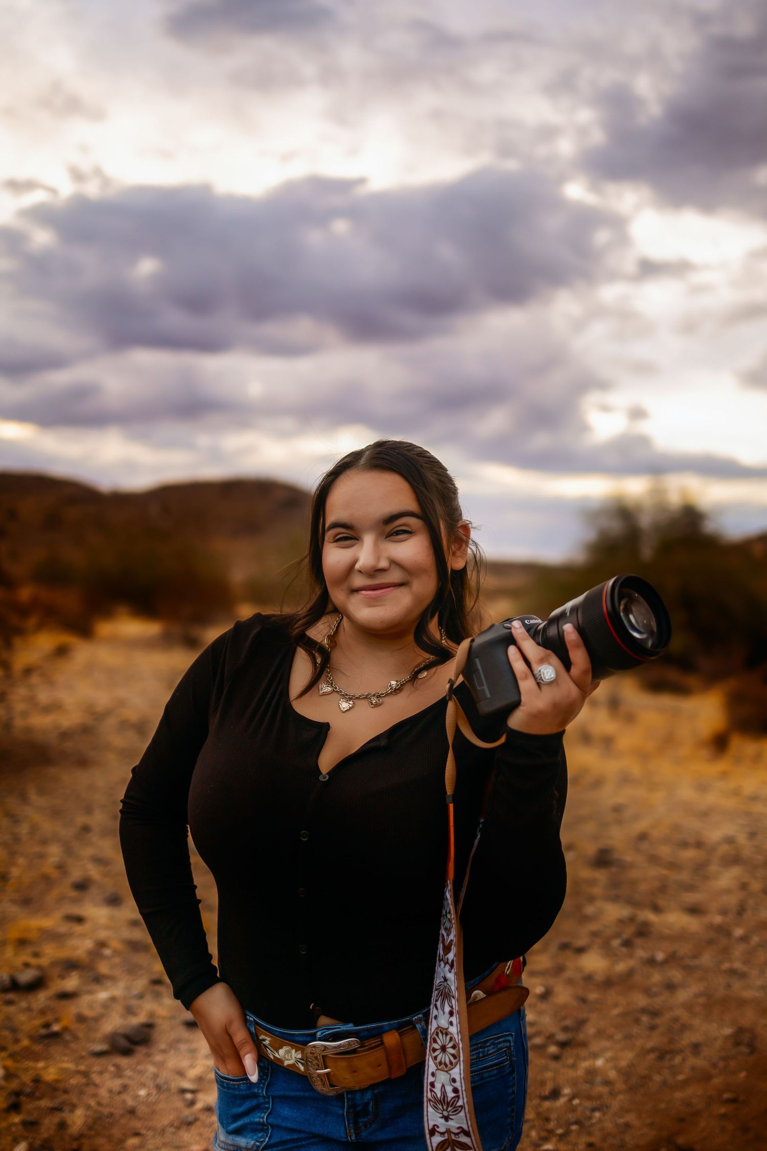 Phoenix Arizona Photographer holding professional camera smiles while standing in desert landscape during golden hour with dramatic clouds.