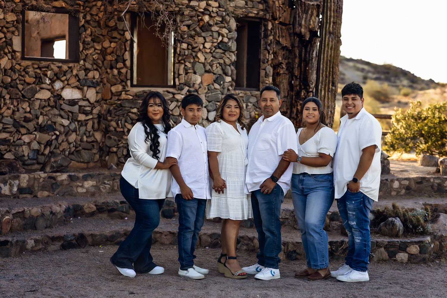 Family group poses together on stone steps in front of rustic stone building in desert landscape at sunset.