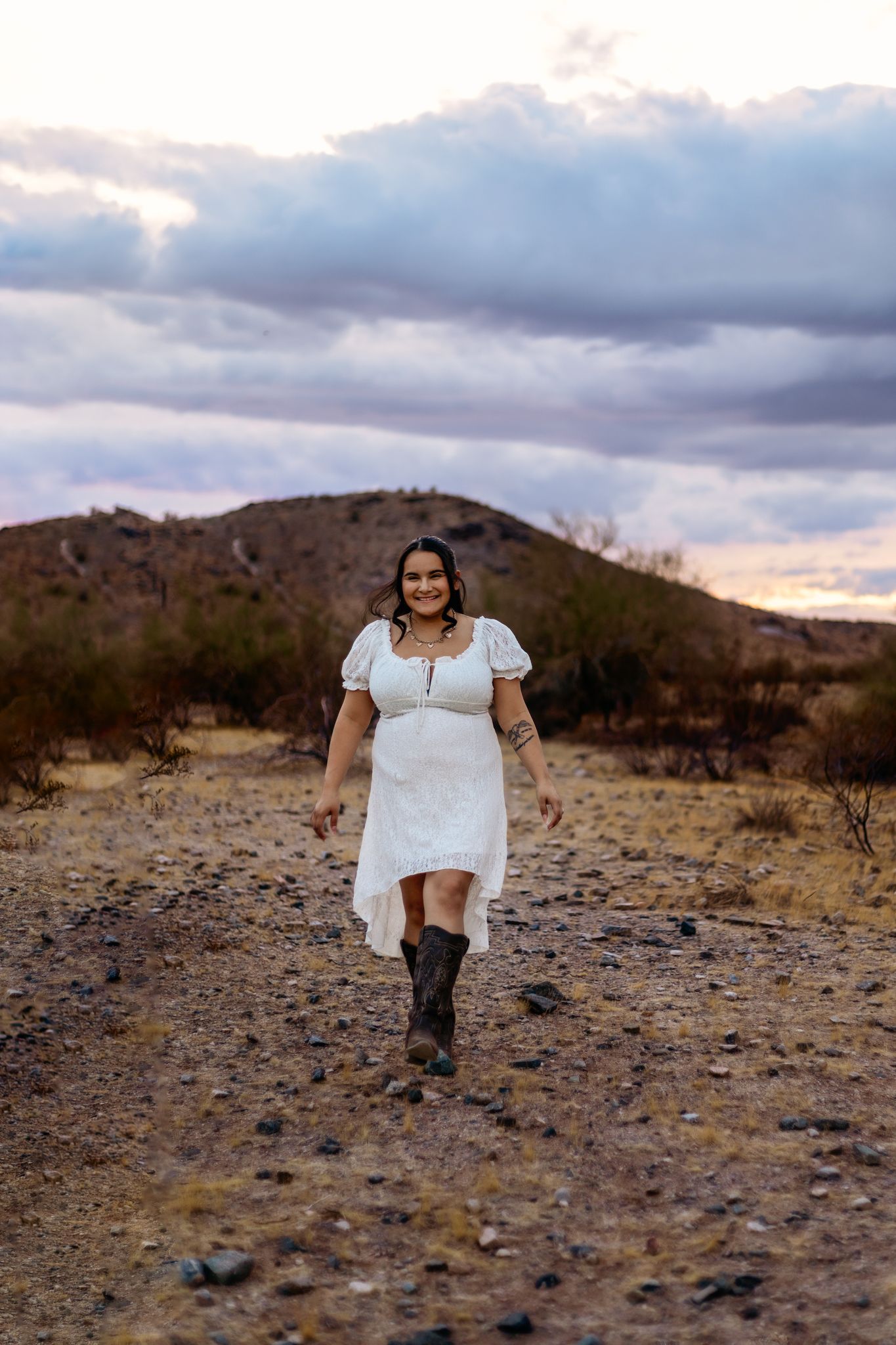 Phoenix Arizona Photographer in a white dress and boots in a desert setting with mountains and dramatic clouds at sunset in the background