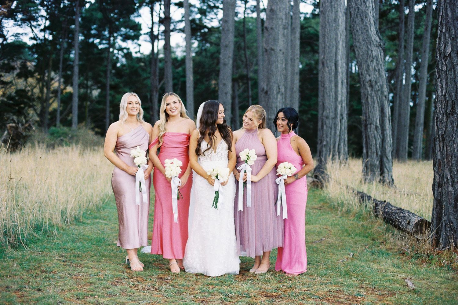 A bridal party wearing different shades of pink and mauve dresses stands together in a forest clearing.
